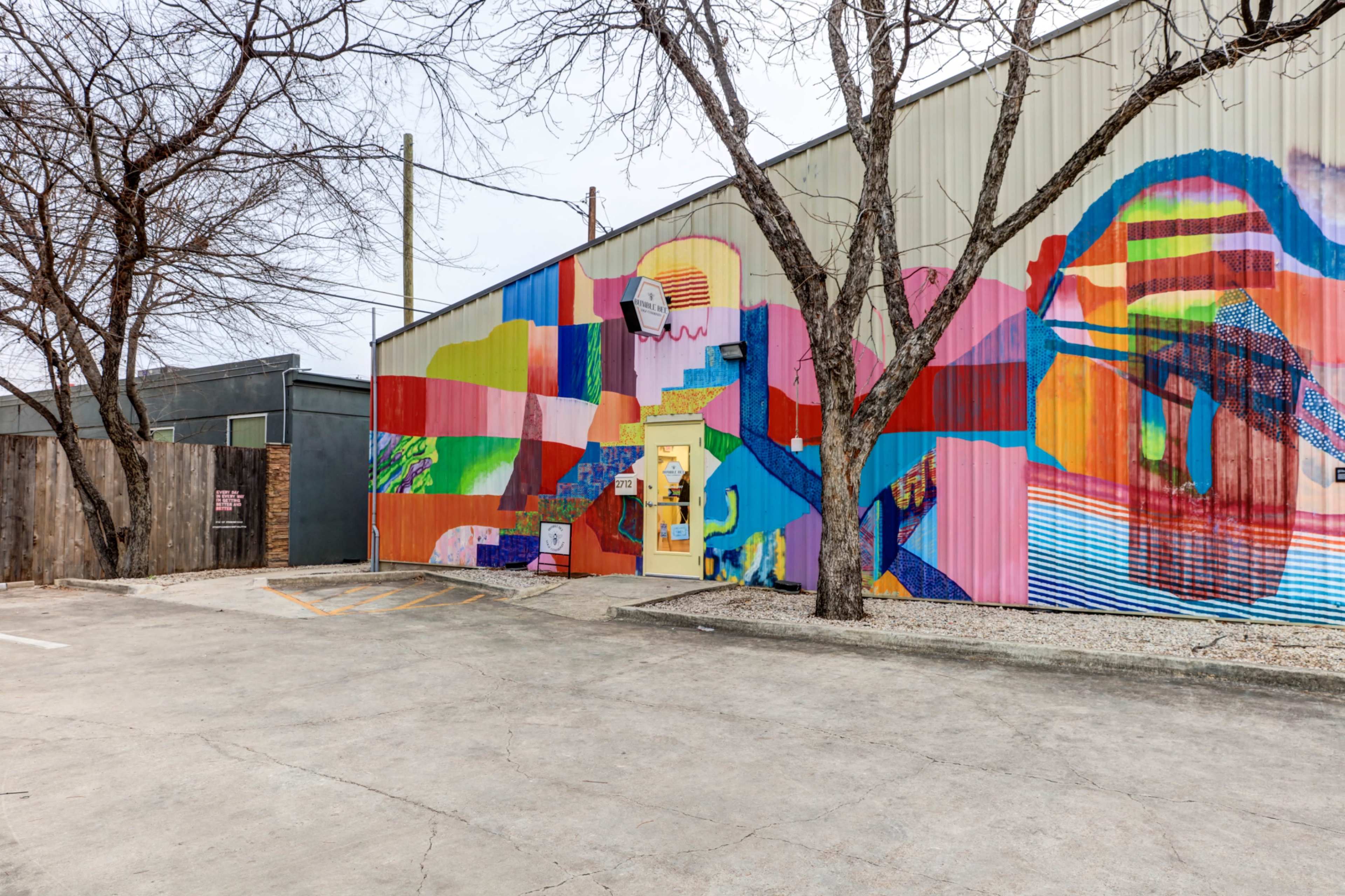 A colorful mural painted on the side of a metal building, with a parking area and bare trees in the foreground.
