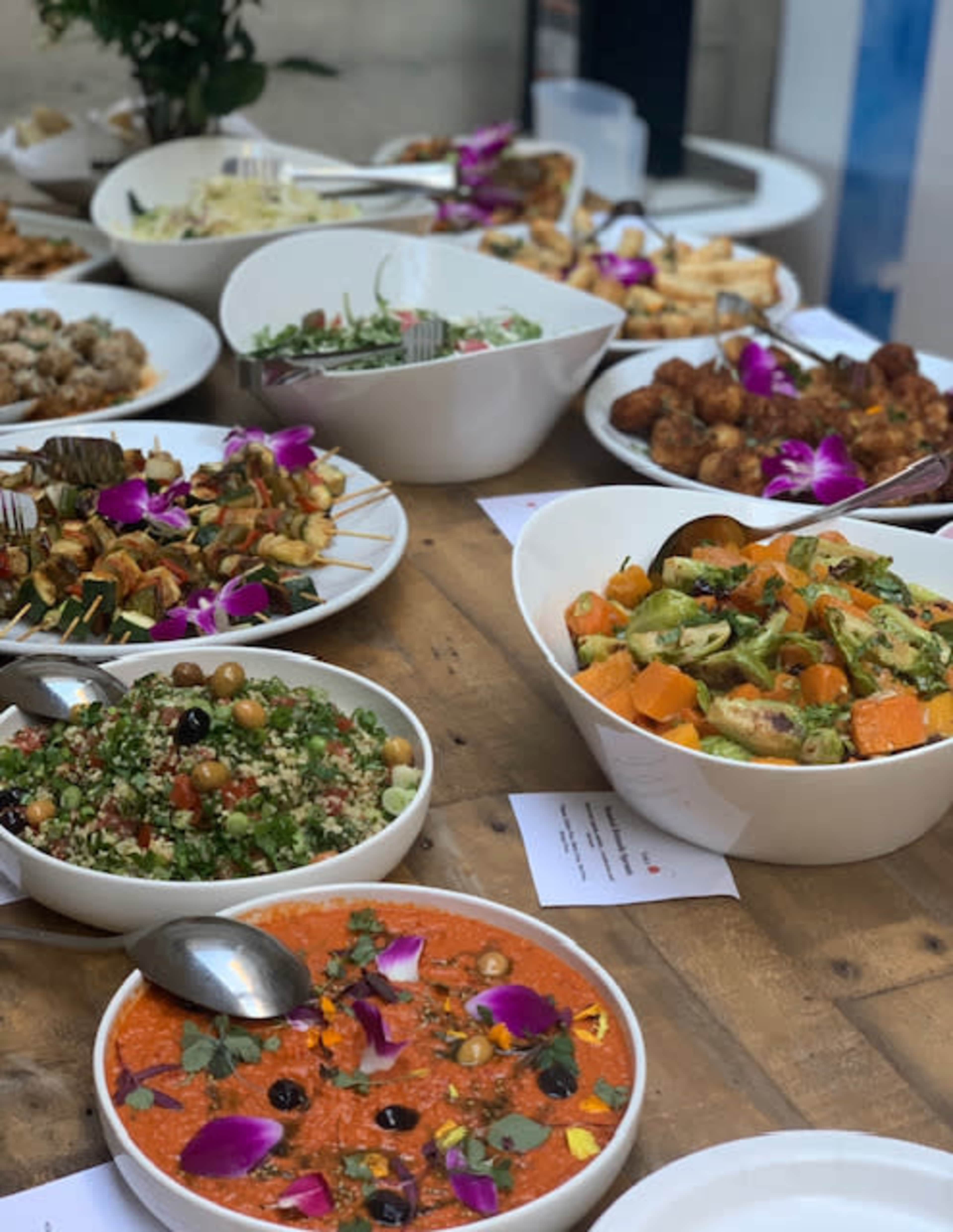 A variety of colorful dishes are arranged on a wooden table, featuring salads, dips, and skewers garnished with flowers.