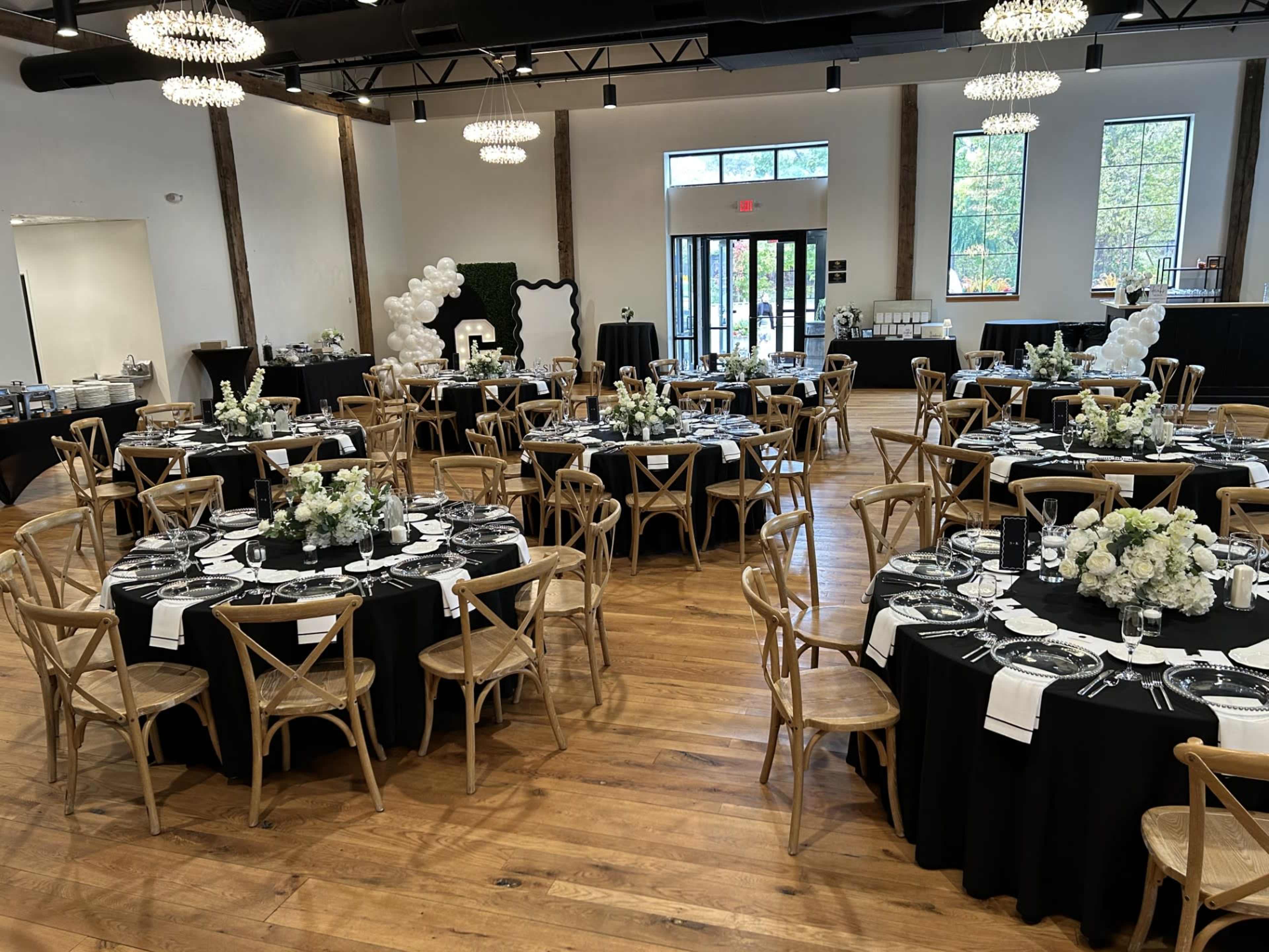 A spacious banquet room is set with round tables adorned with black tablecloths and white floral centerpieces, while chandeliers hang above.