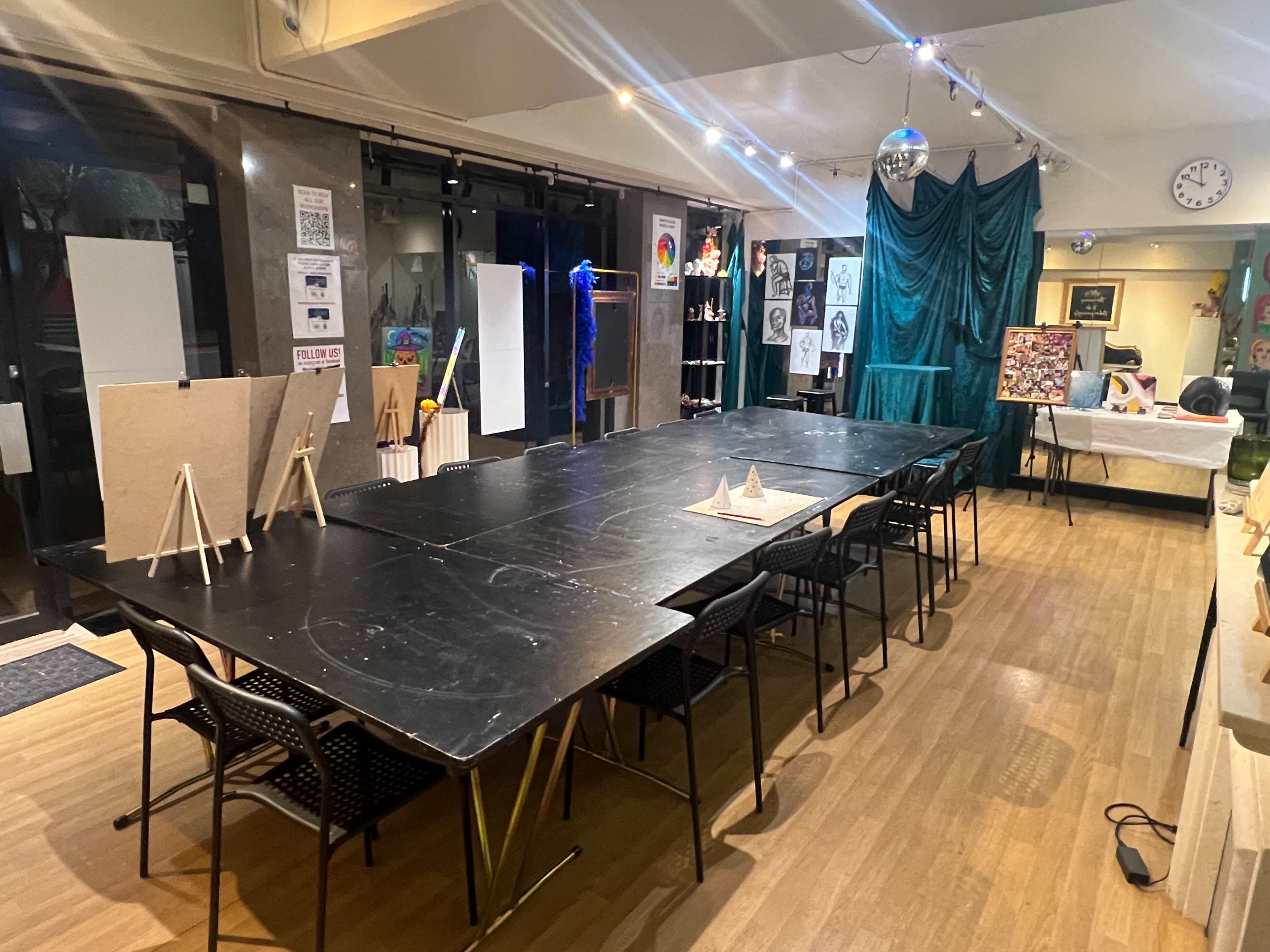 The image shows an art studio with a long black table surrounded by chairs, several easels displayed along the walls, and various artwork pieces positioned throughout the space.