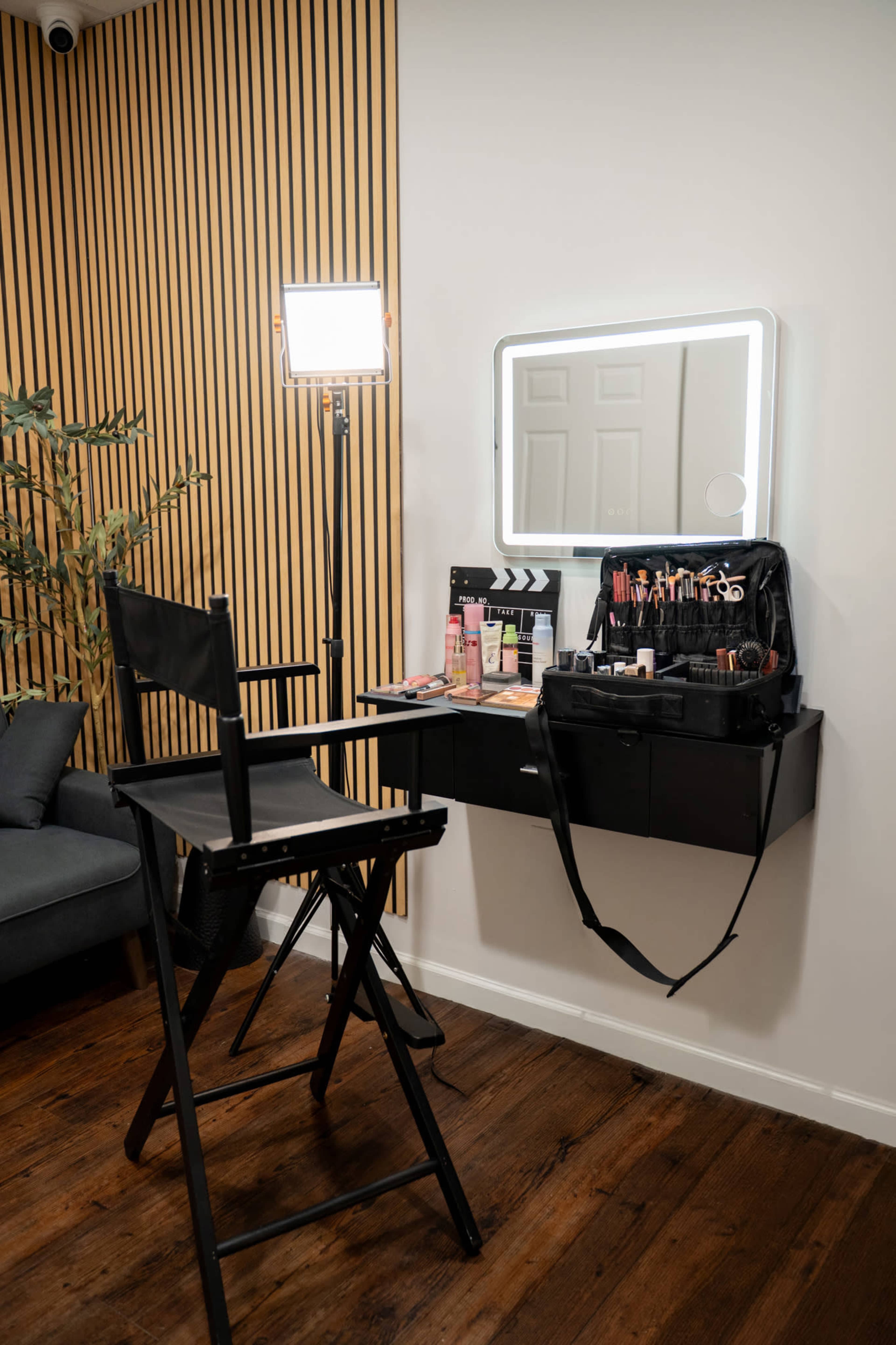 The image shows a makeup station with a tall director's chair, a wall-mounted mirror, and a neatly organized makeup kit and products on a shelf, set against a backdrop of wooden strips.
