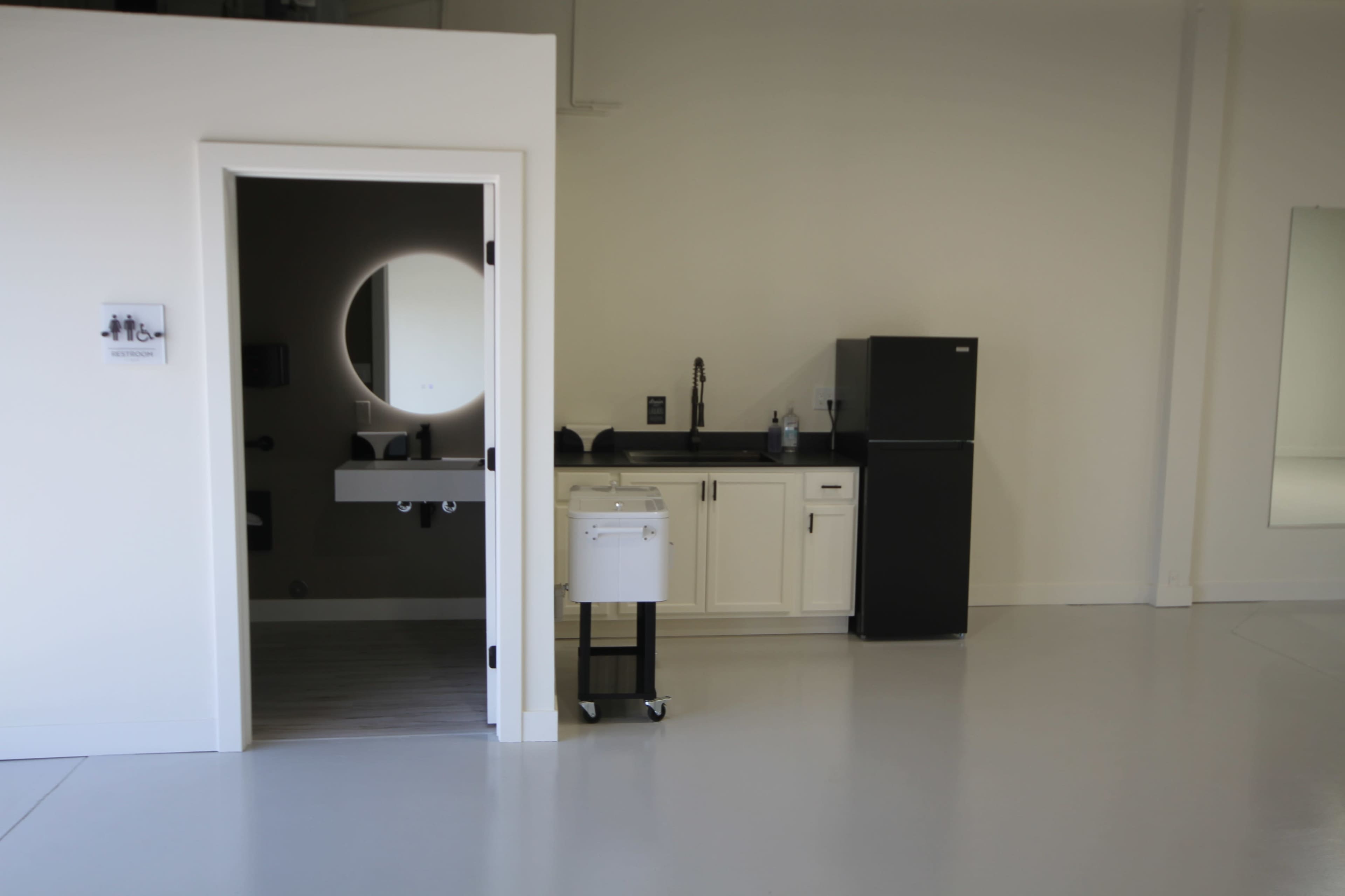 The image shows a spacious room with a small kitchenette, a bathroom area with a round mirror, and a black refrigerator.