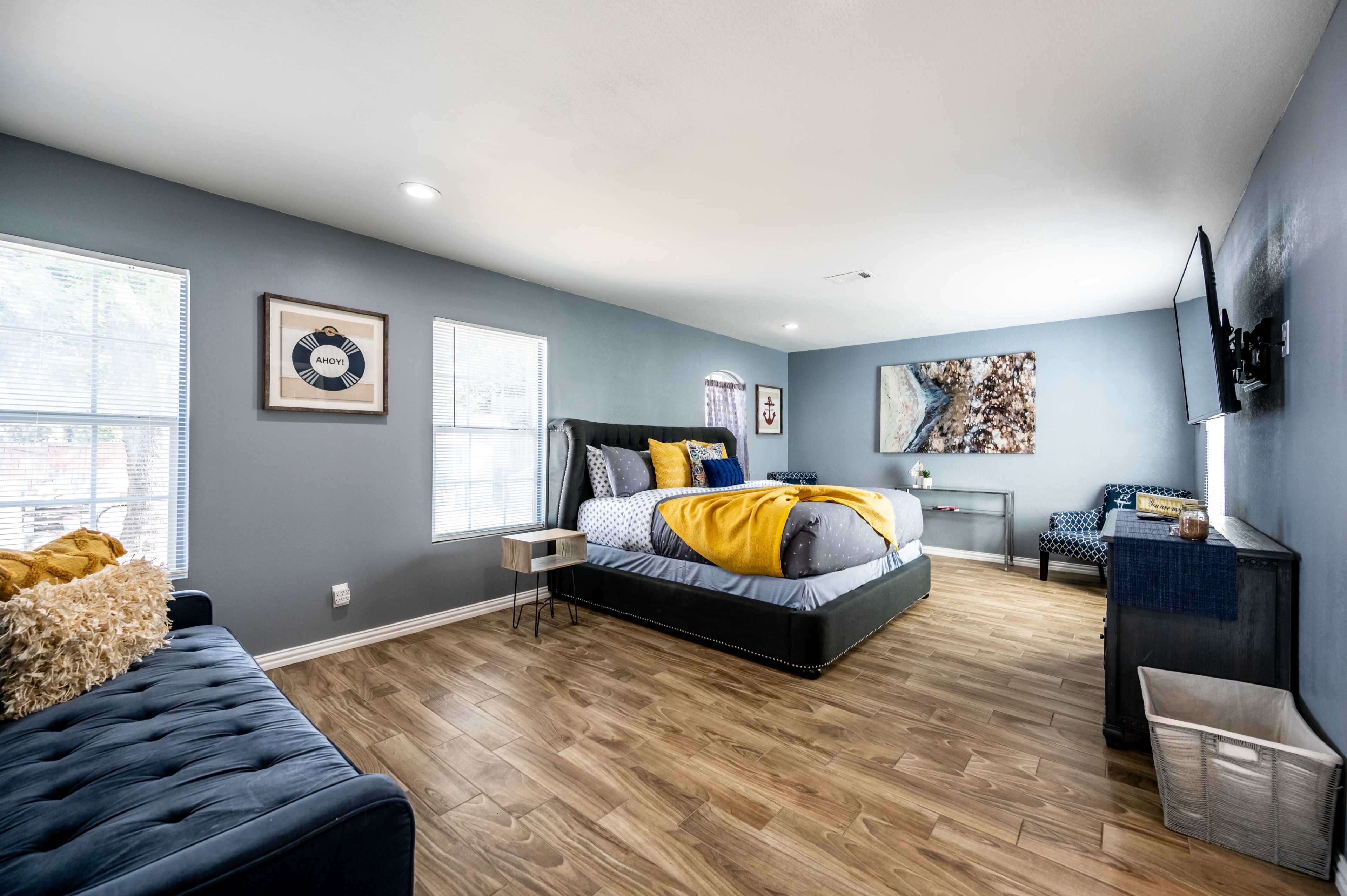The room features a bed with yellow and gray bedding, a blue sofa, and a television mounted on the wall, with windows allowing natural light to enter.