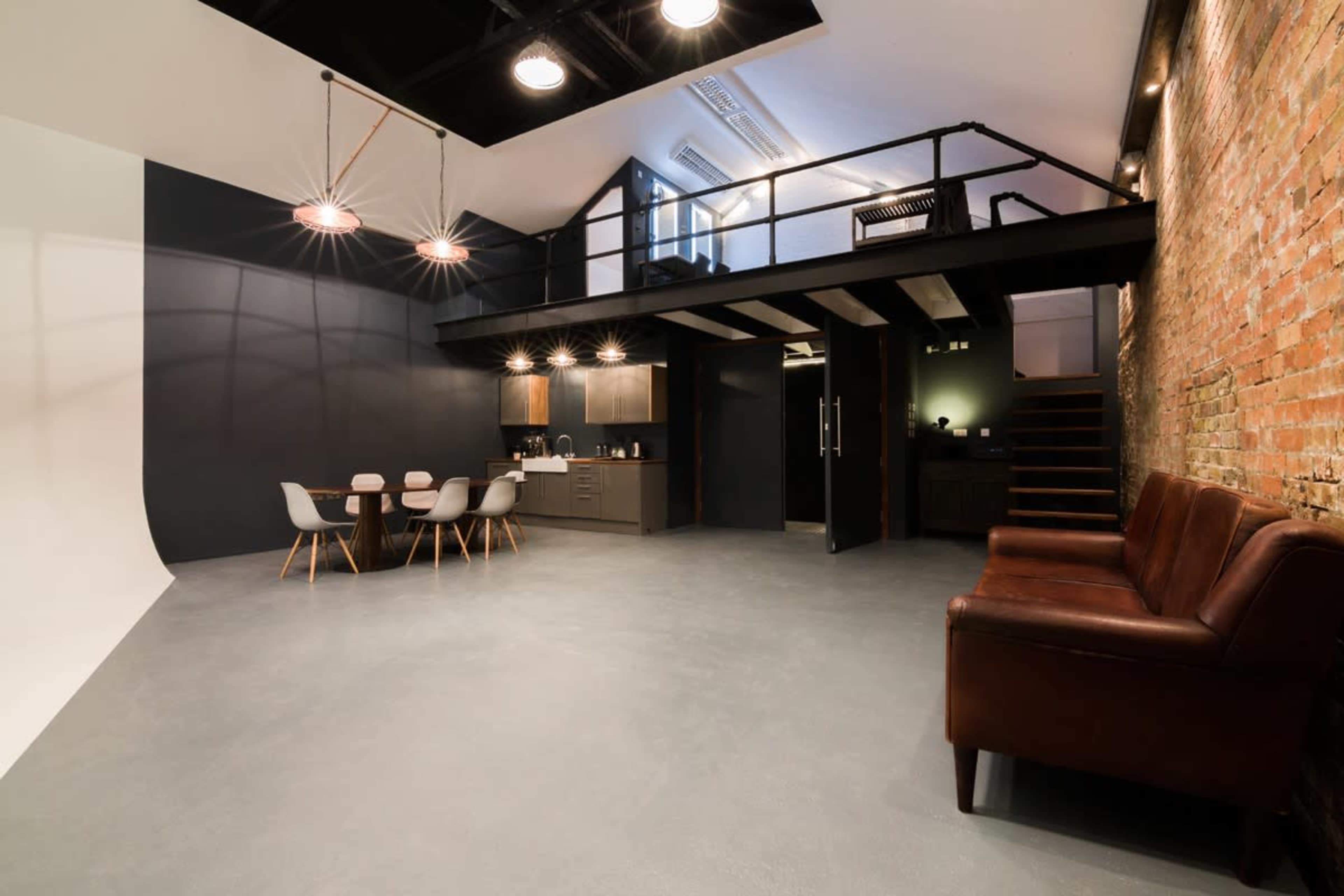The image depicts a modern studio space featuring a lofted area, a kitchen, a dining table with chairs, a leather couch, and exposed brick walls.