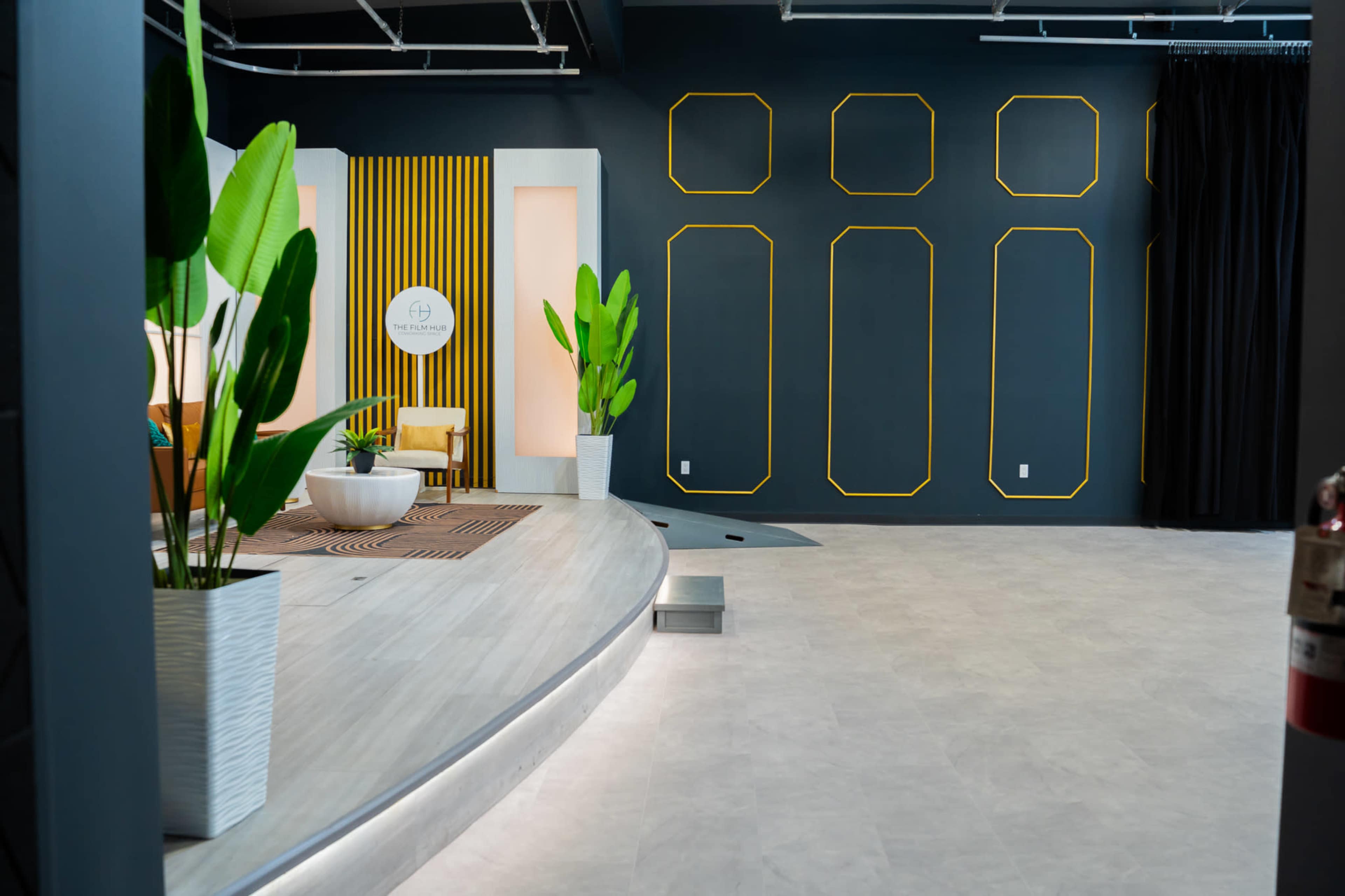 The image shows a modern interior space with a round, raised platform, a geometric wall design in dark colors, and potted plants in white containers.