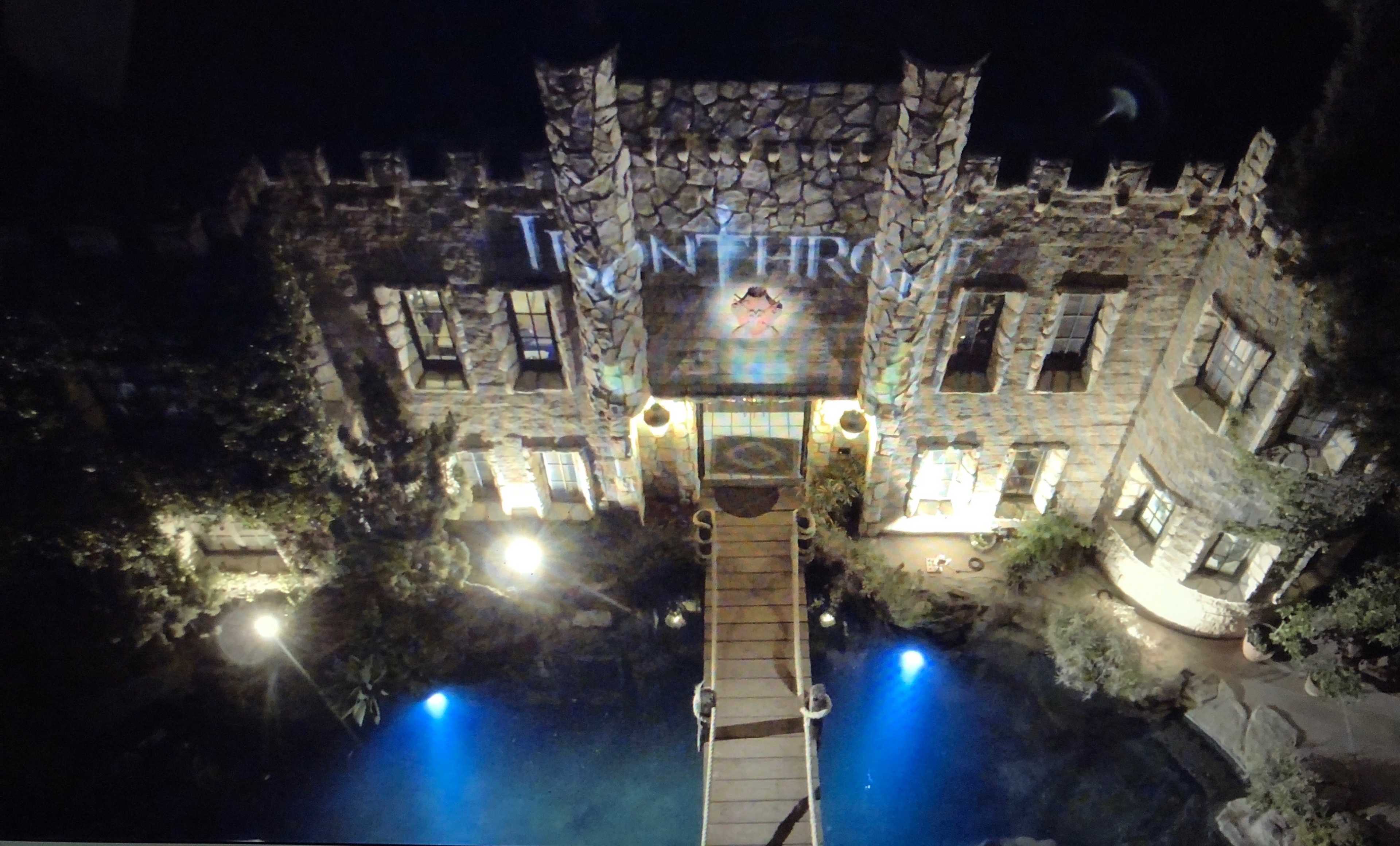 A stone mansion with a bridge over a water feature is illuminated at night, featuring a projection on its facade.