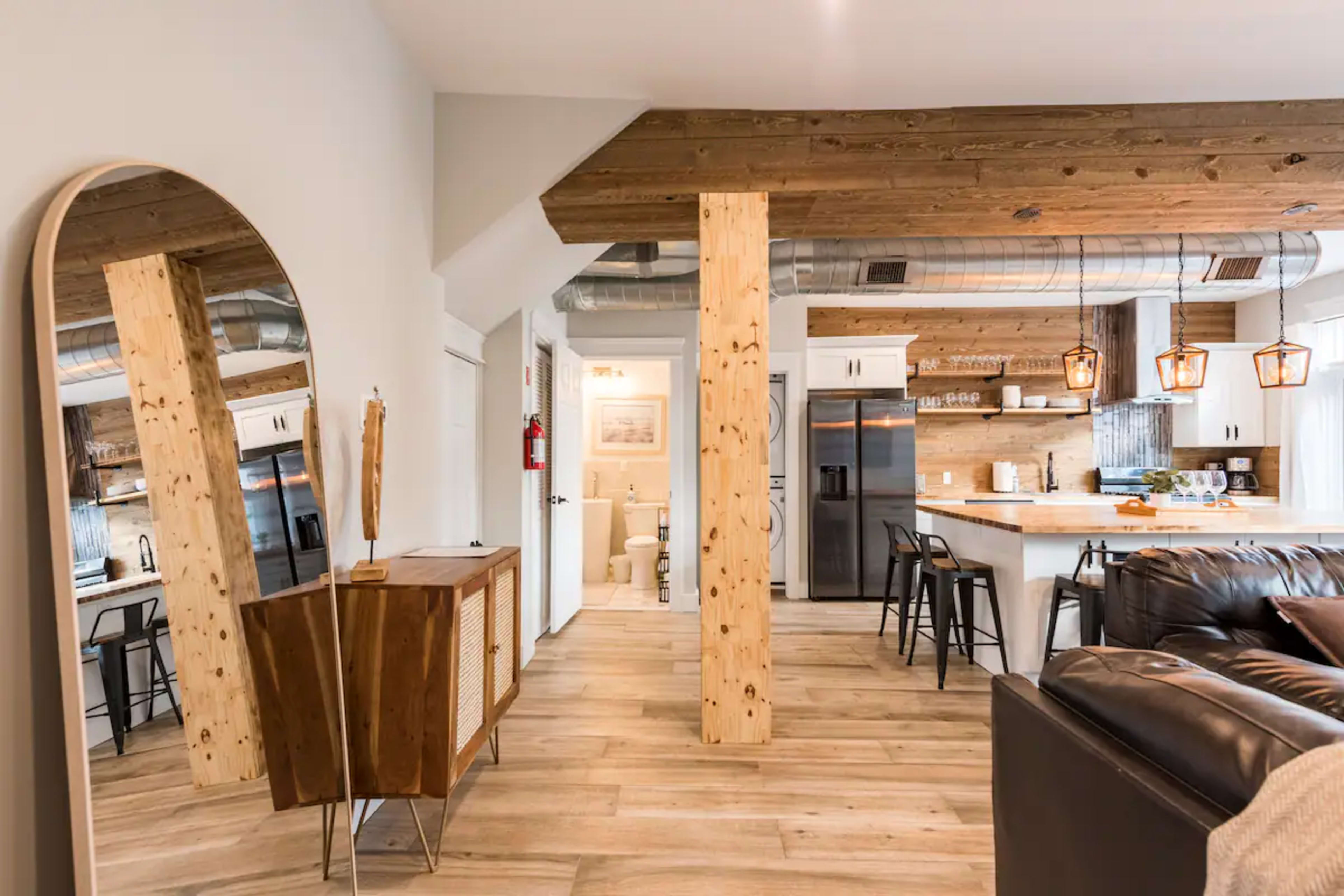 The image shows a modern living space featuring an open layout with a kitchen, dining area, and living room, characterized by wooden accents and stylish furnishings.