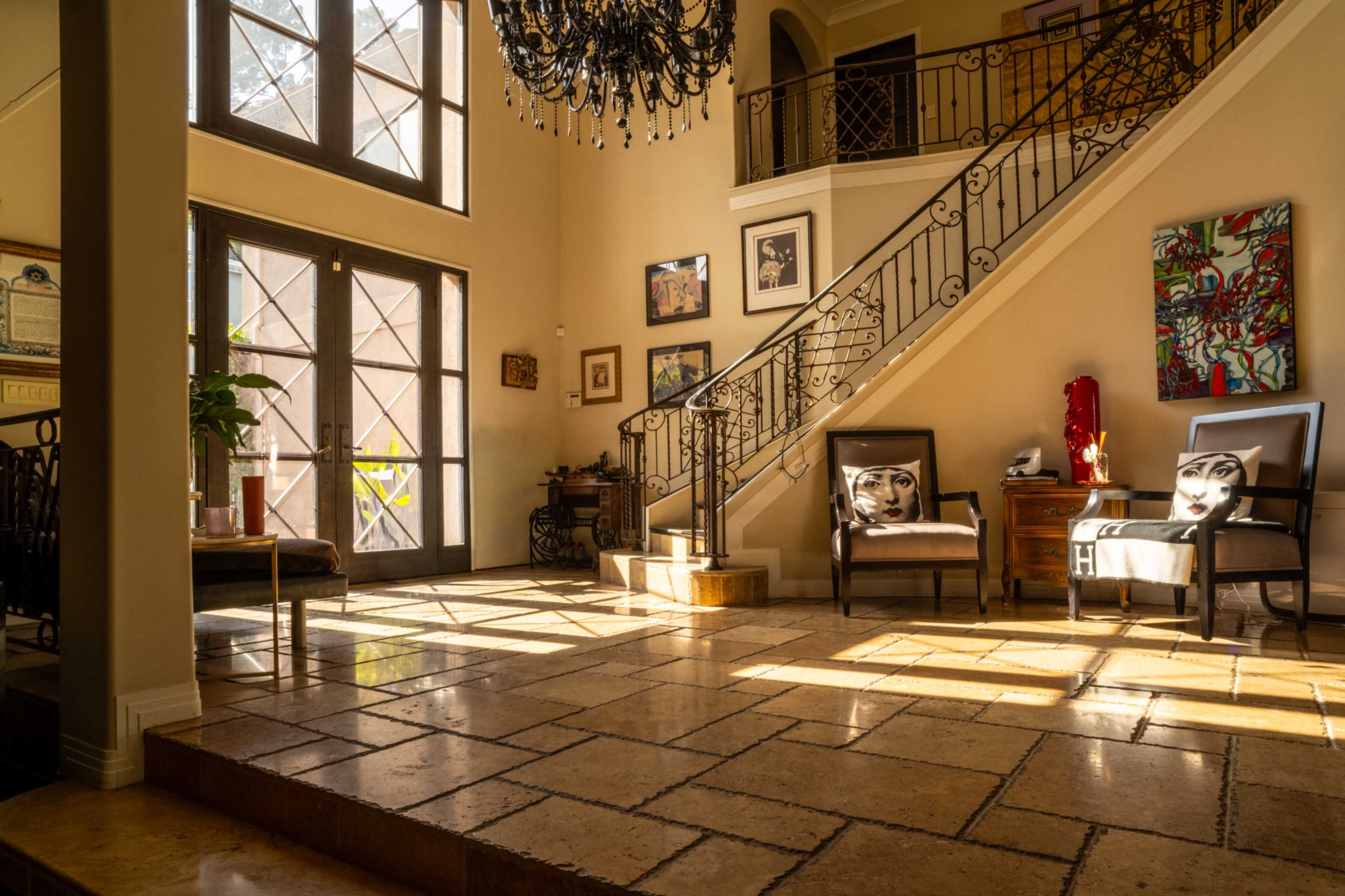The image shows a spacious entryway with a large chandelier, a staircase, two chairs, and artwork displayed on the walls.