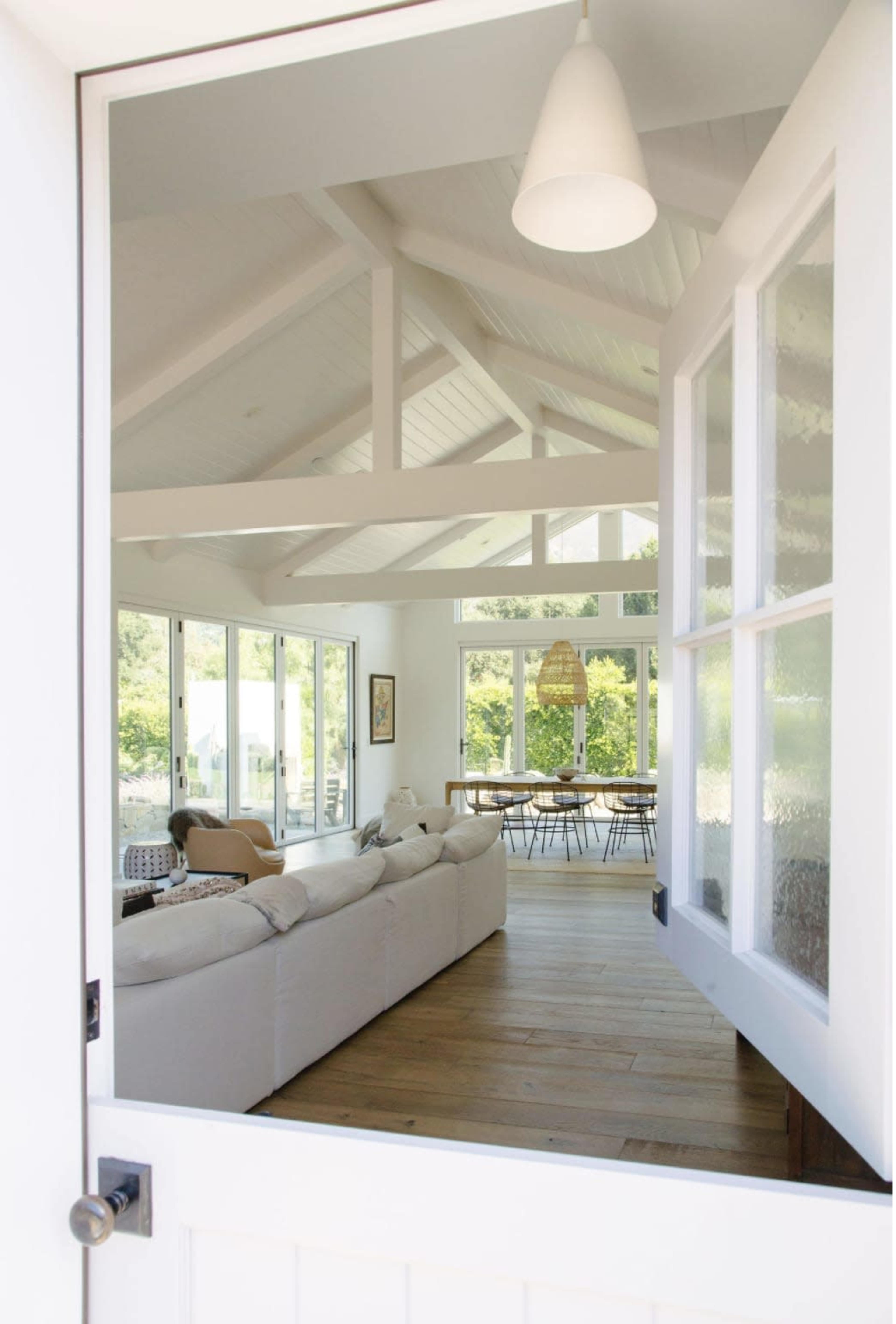 A modern, open living space is visible through a doorway, featuring a large sofa, wooden dining table, and expansive windows that let in natural light.