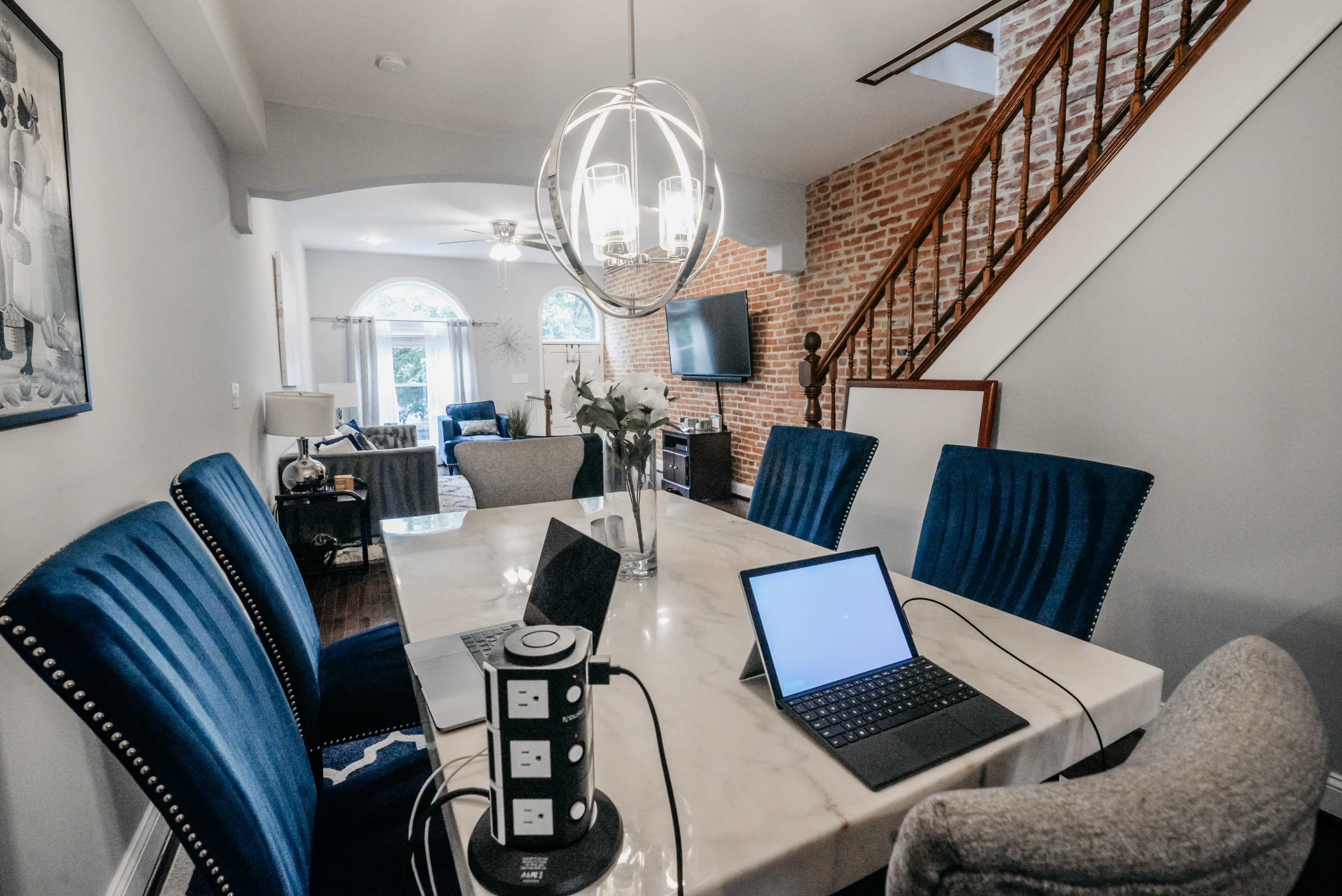 A modern dining area features a marble table with blue upholstered chairs, surrounded by a staircase and a living space in the background.