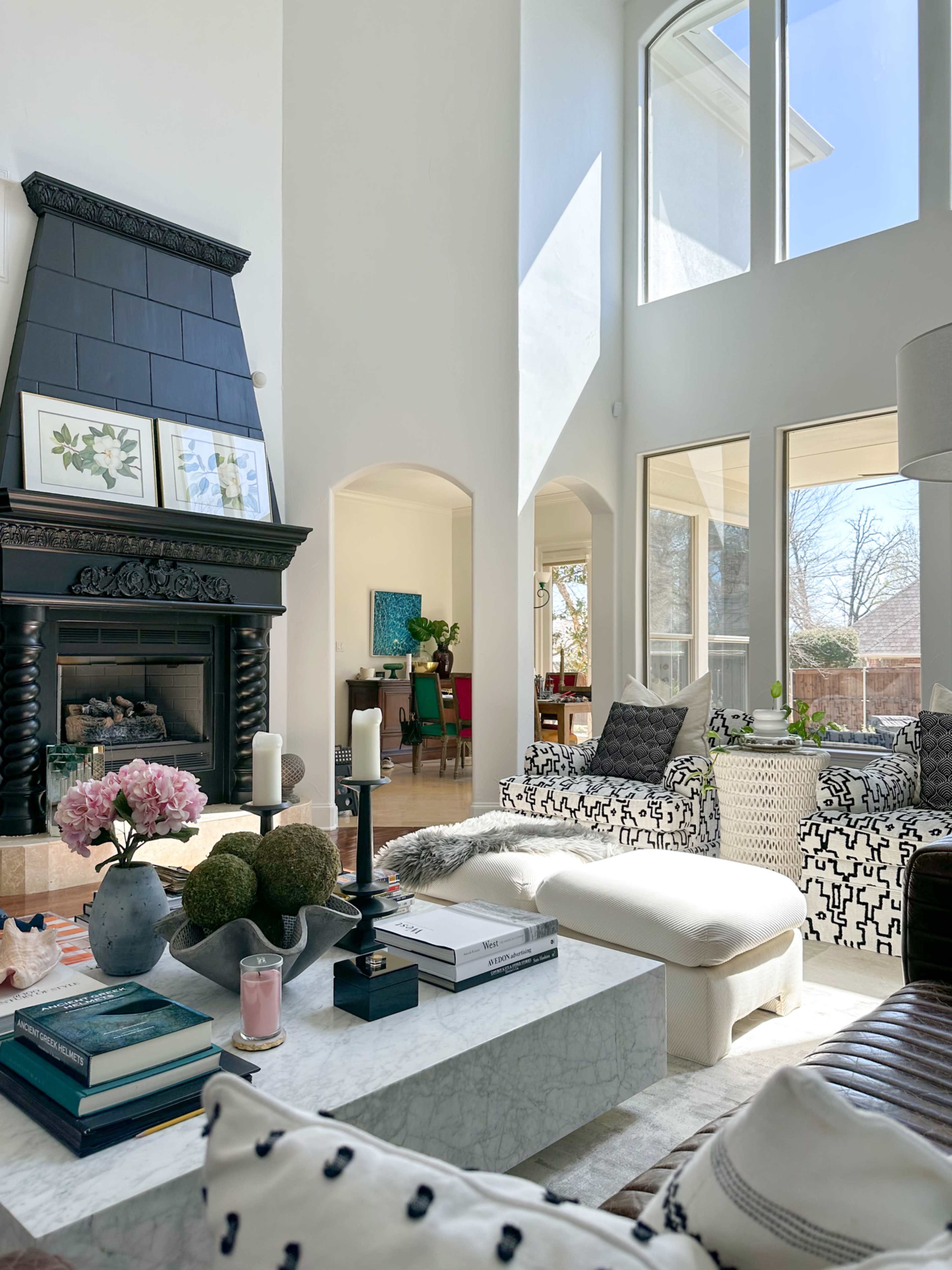 The bright living room features high ceilings, a black fireplace, and a mix of patterned furniture surrounding a coffee table on a textured rug.