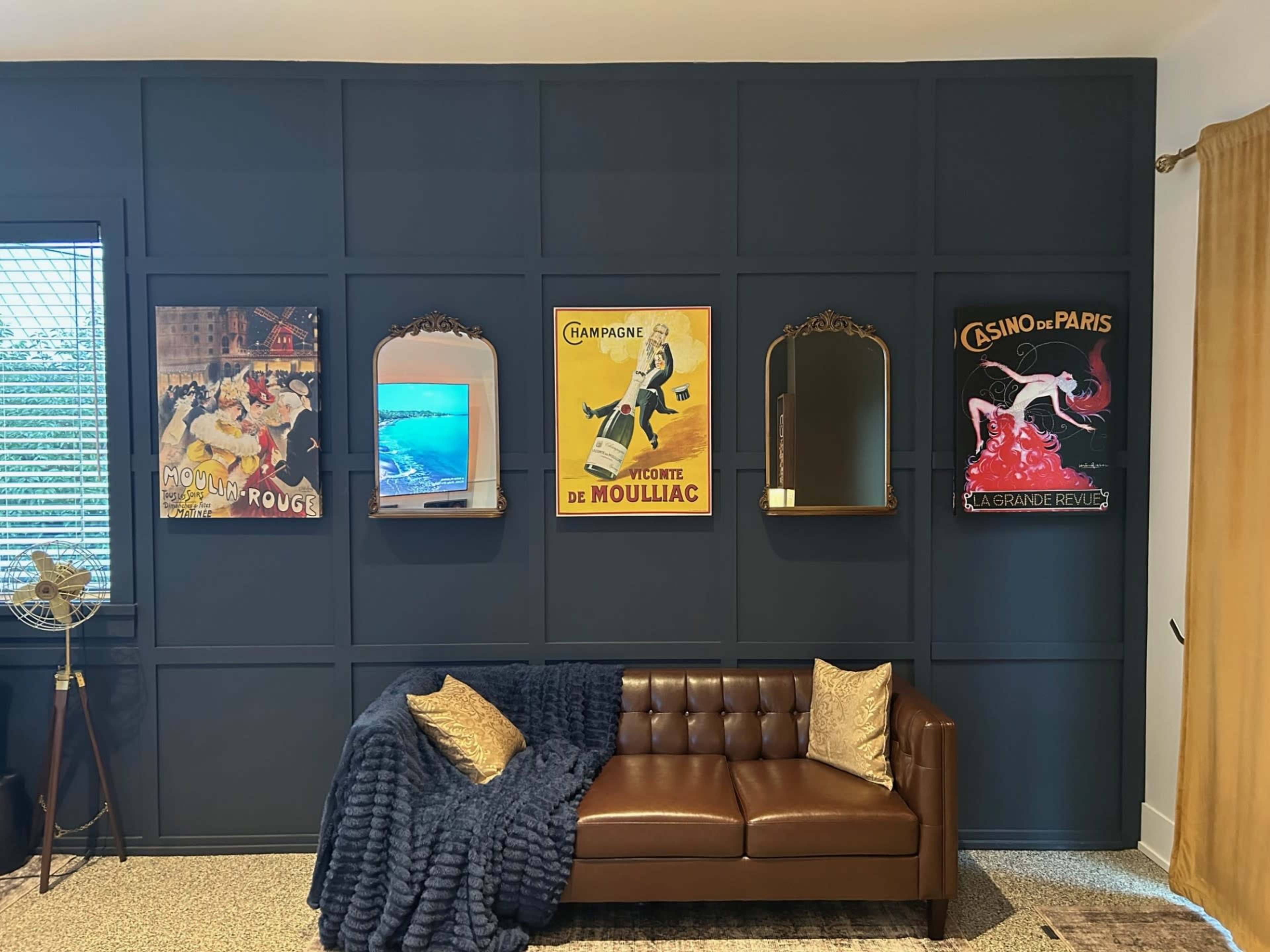 The image features a dark blue accent wall adorned with vintage-style posters, a decorative mirror, and a brown leather couch draped with a blue throw blanket.