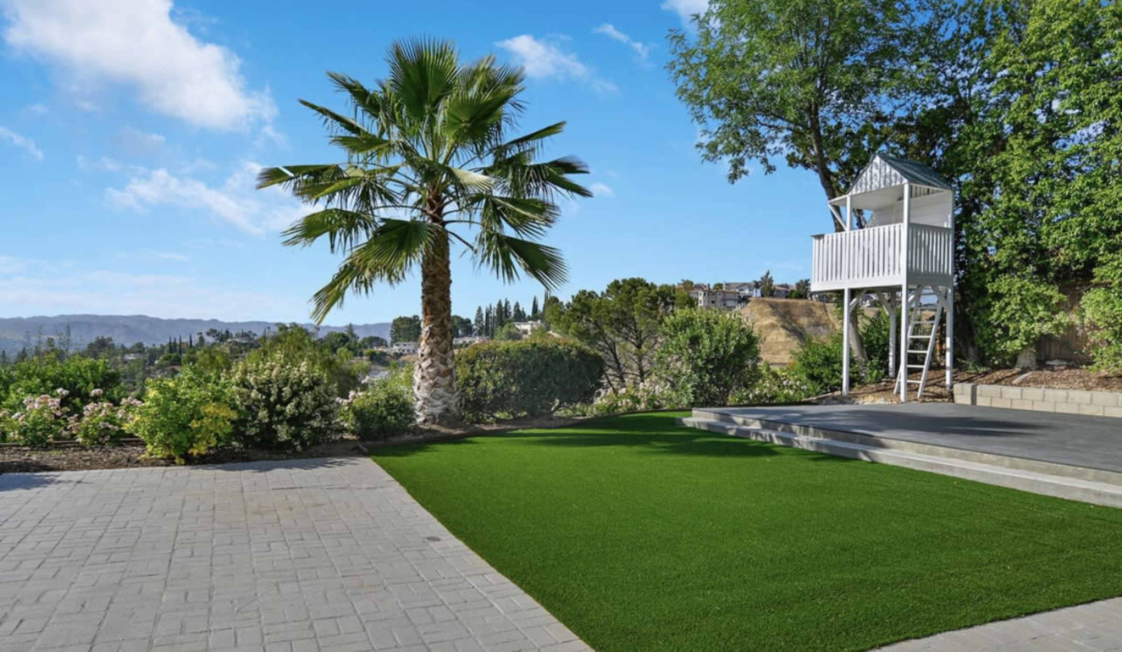 A landscaped yard features a palm tree, a small playhouse, and a patch of green grass surrounded by garden areas.