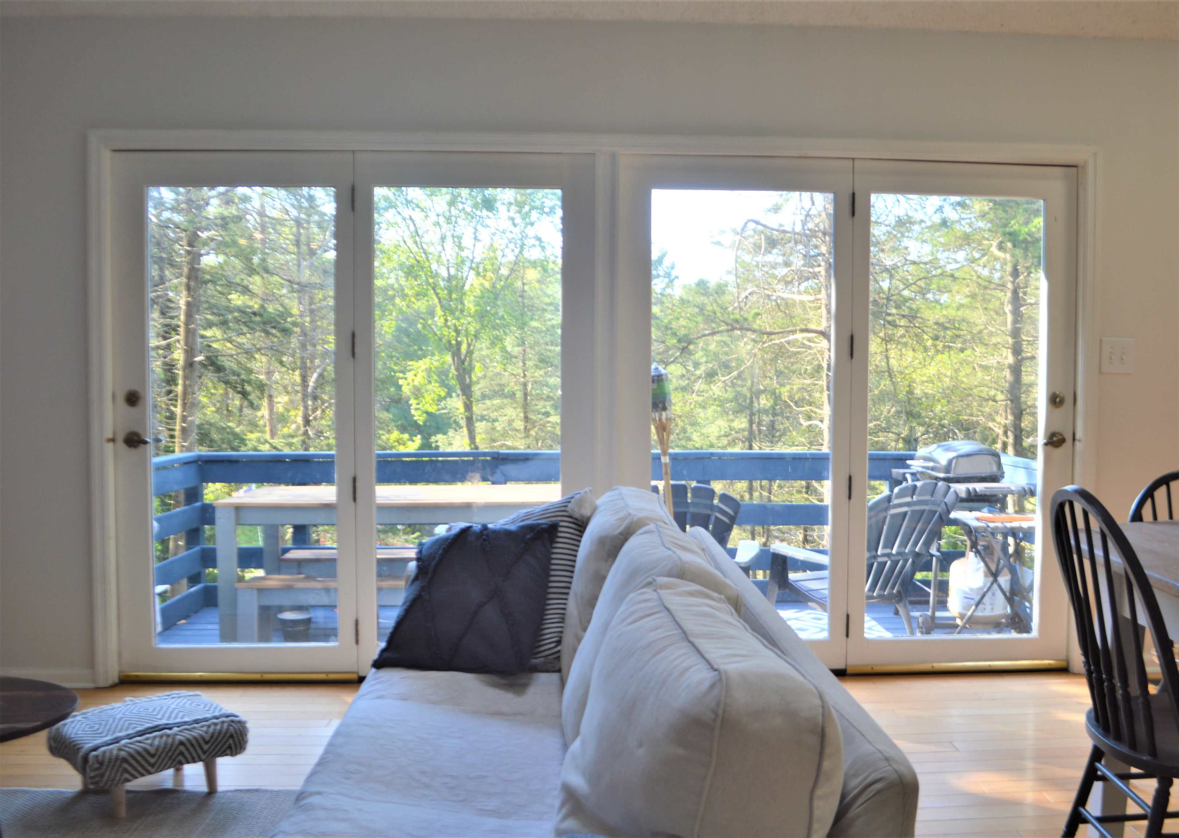 A living room with a light-colored sofa facing a large set of glass doors that open to a deck surrounded by trees.