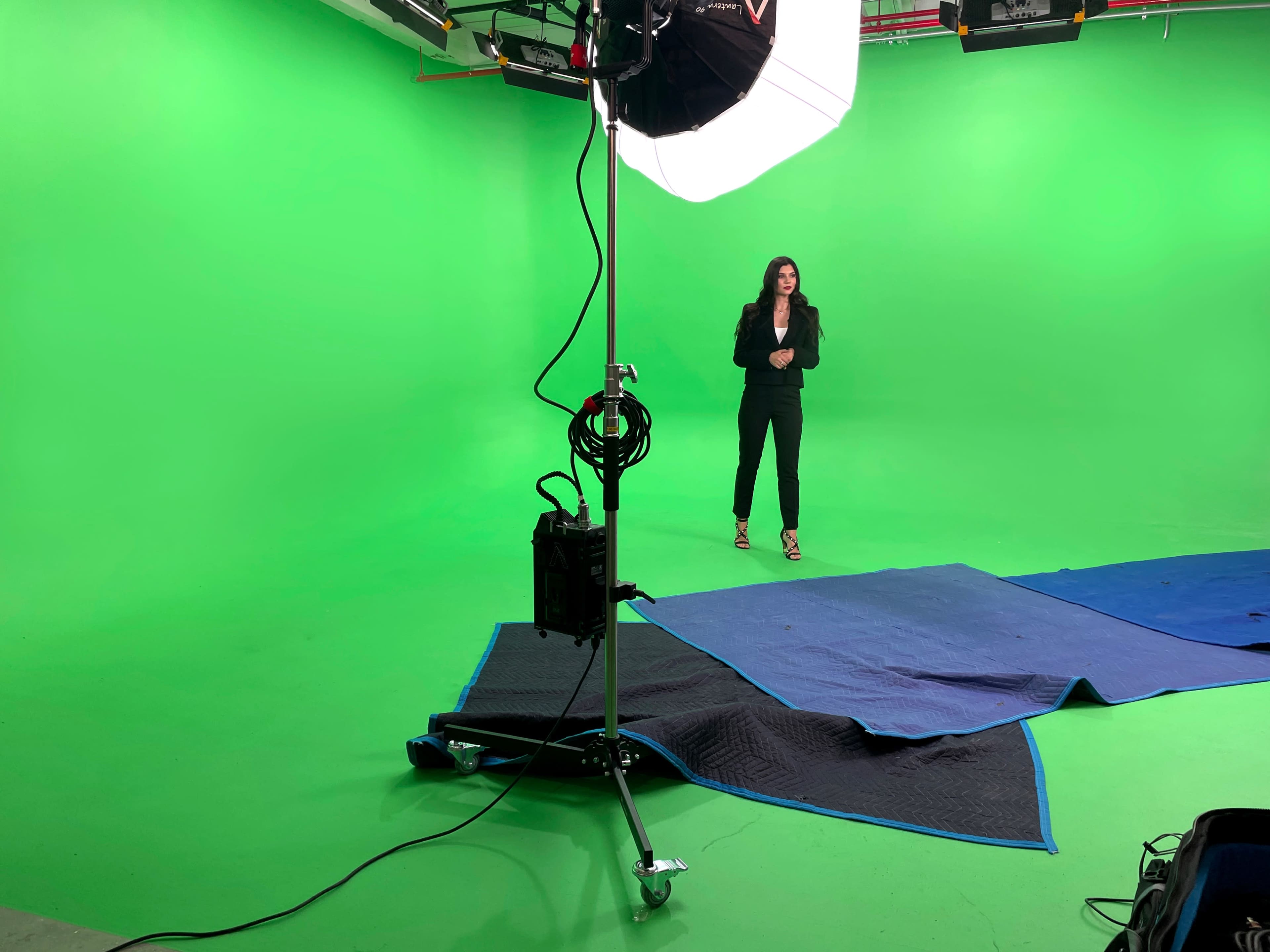A person stands in front of a green screen in a studio setting, with lighting equipment and protective mats on the floor.
