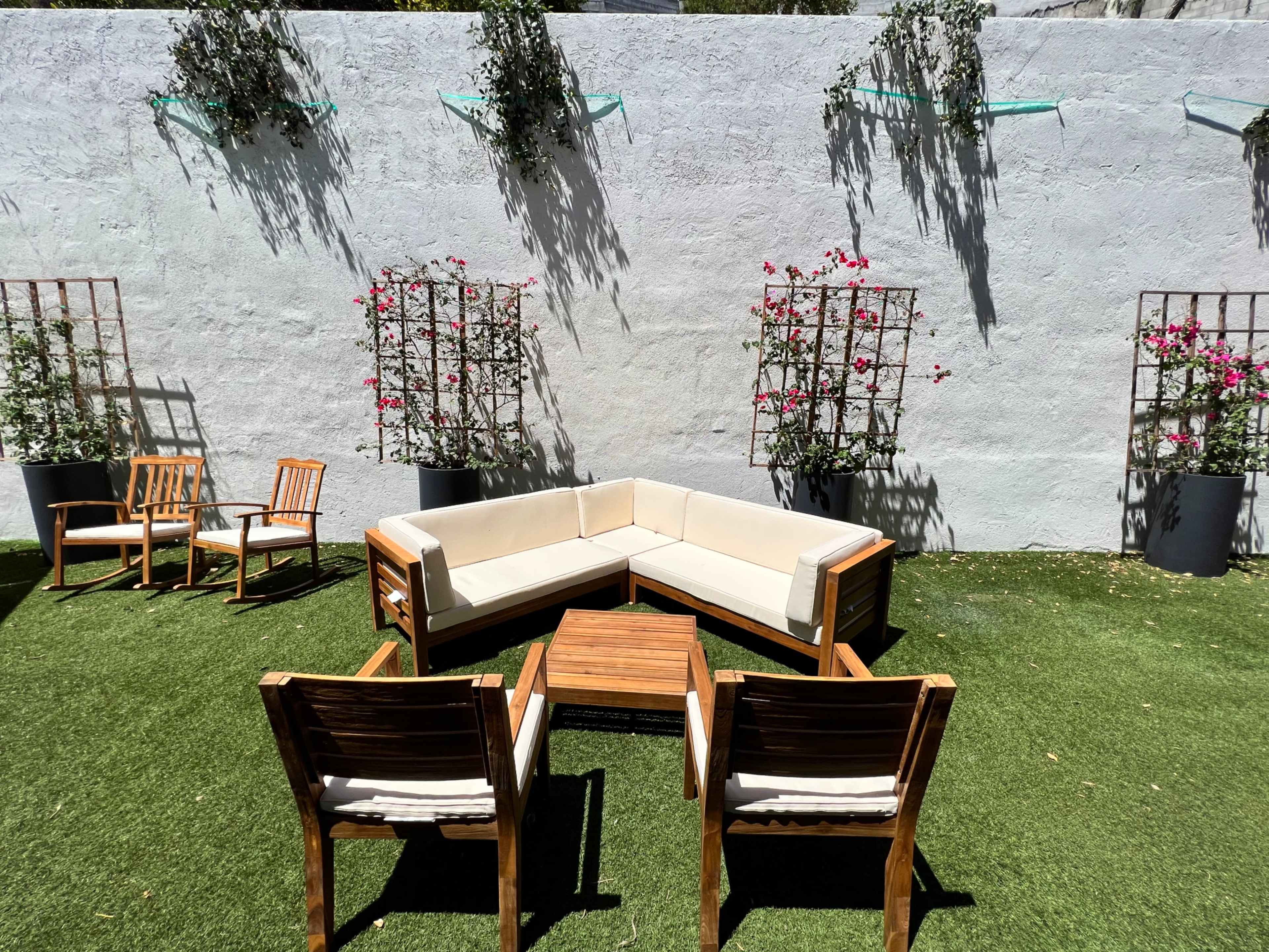 A landscaped outdoor seating area features a beige sectional sofa, wooden chairs, and a coffee table surrounded by floral planters against a light gray wall.