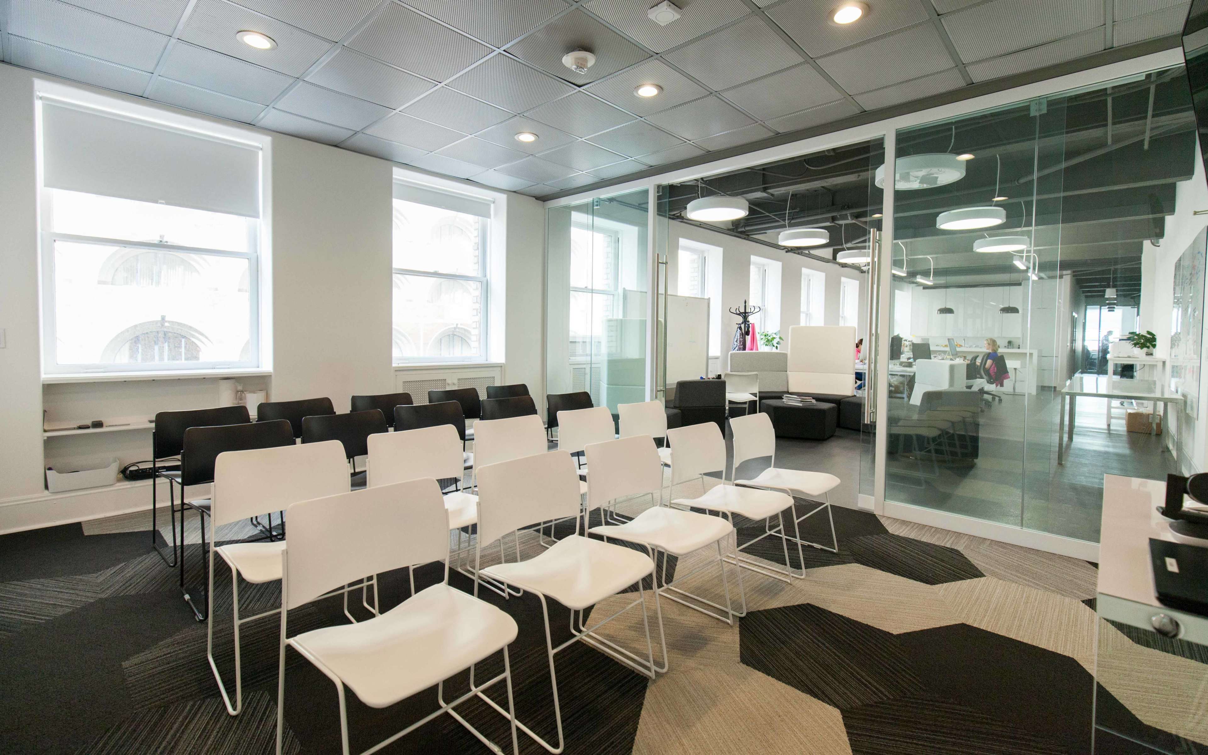 A meeting room features a row of white and black chairs facing a glass partition, with large windows providing natural light.