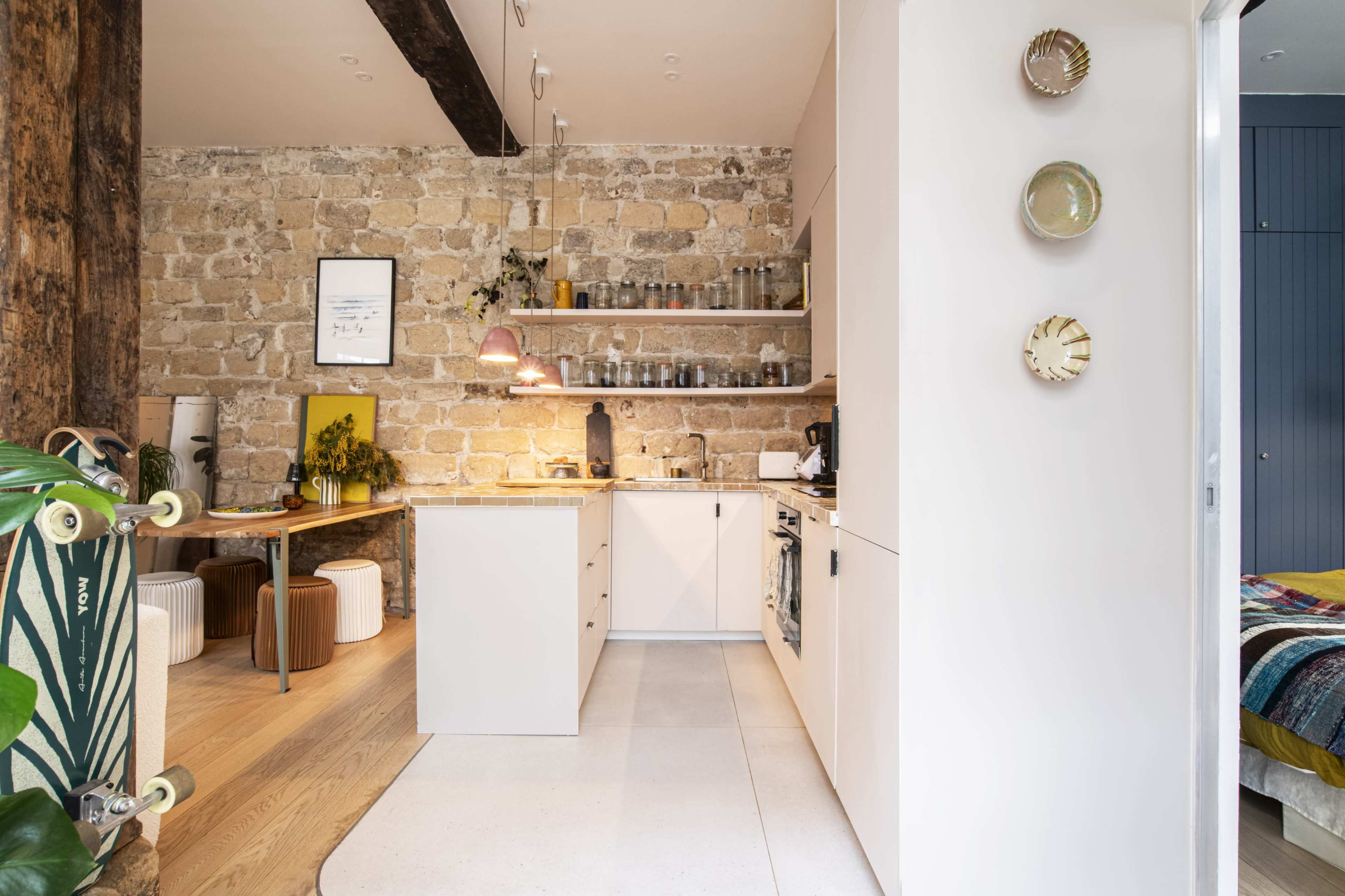 The image shows a modern kitchen with a stone wall, wooden dining table, and open shelving displaying various kitchenware.