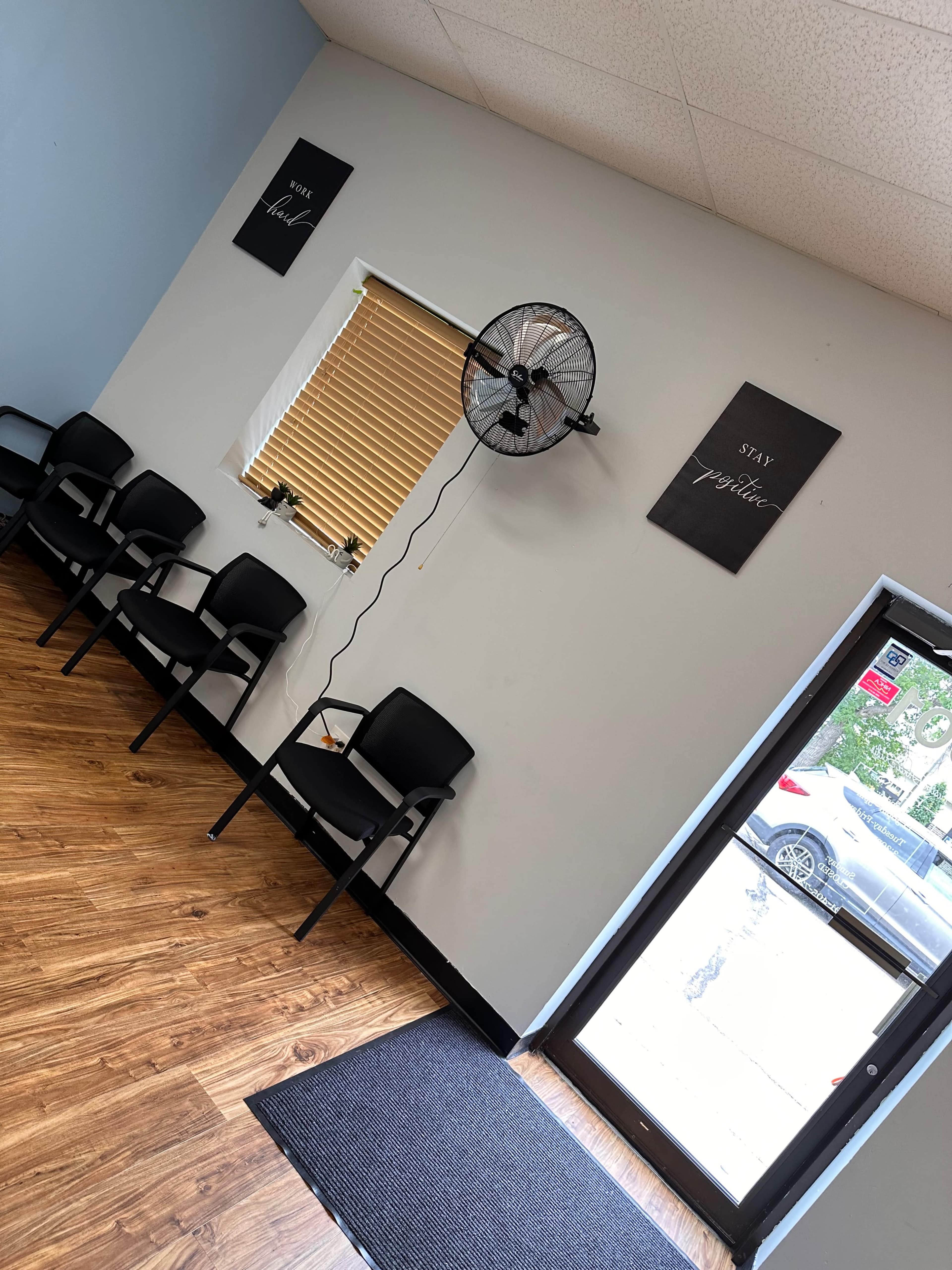 The image shows a waiting area with several black chairs, a wall-mounted fan, and a door leading outside, along with motivational signs on the walls.