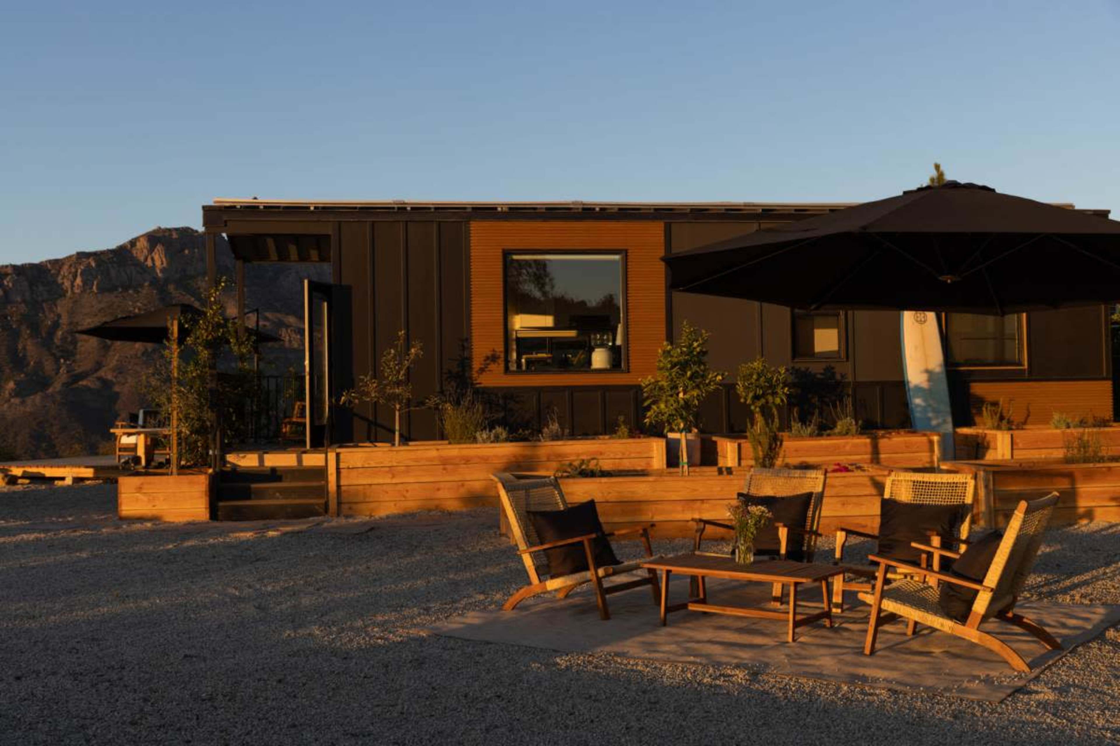 A modern cabin with a wooden deck and outdoor seating area is set against a mountainous backdrop during sunset.