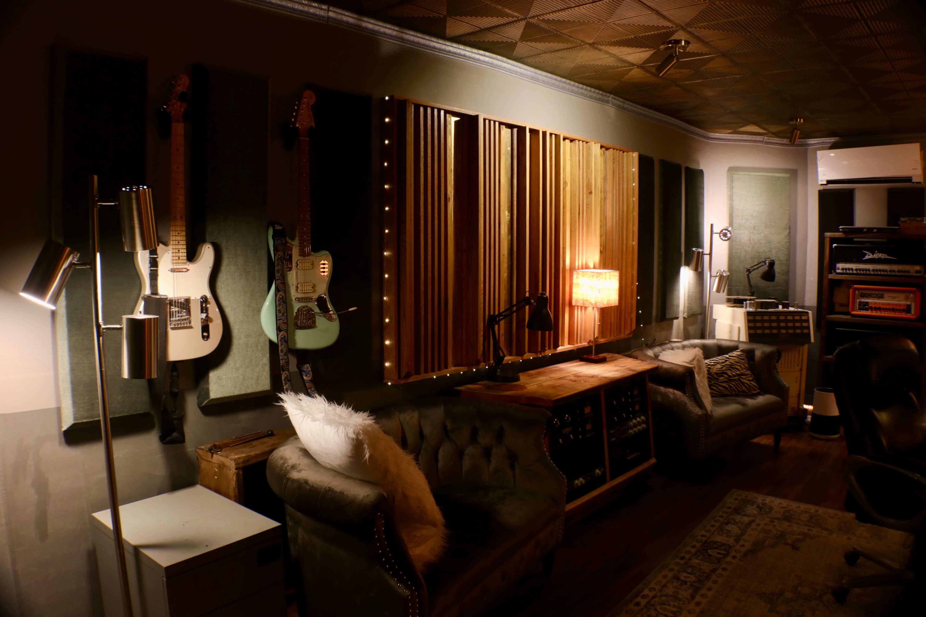 A dimly lit music studio featuring two guitars on the wall, a wooden panel with lights, and comfortable seating.