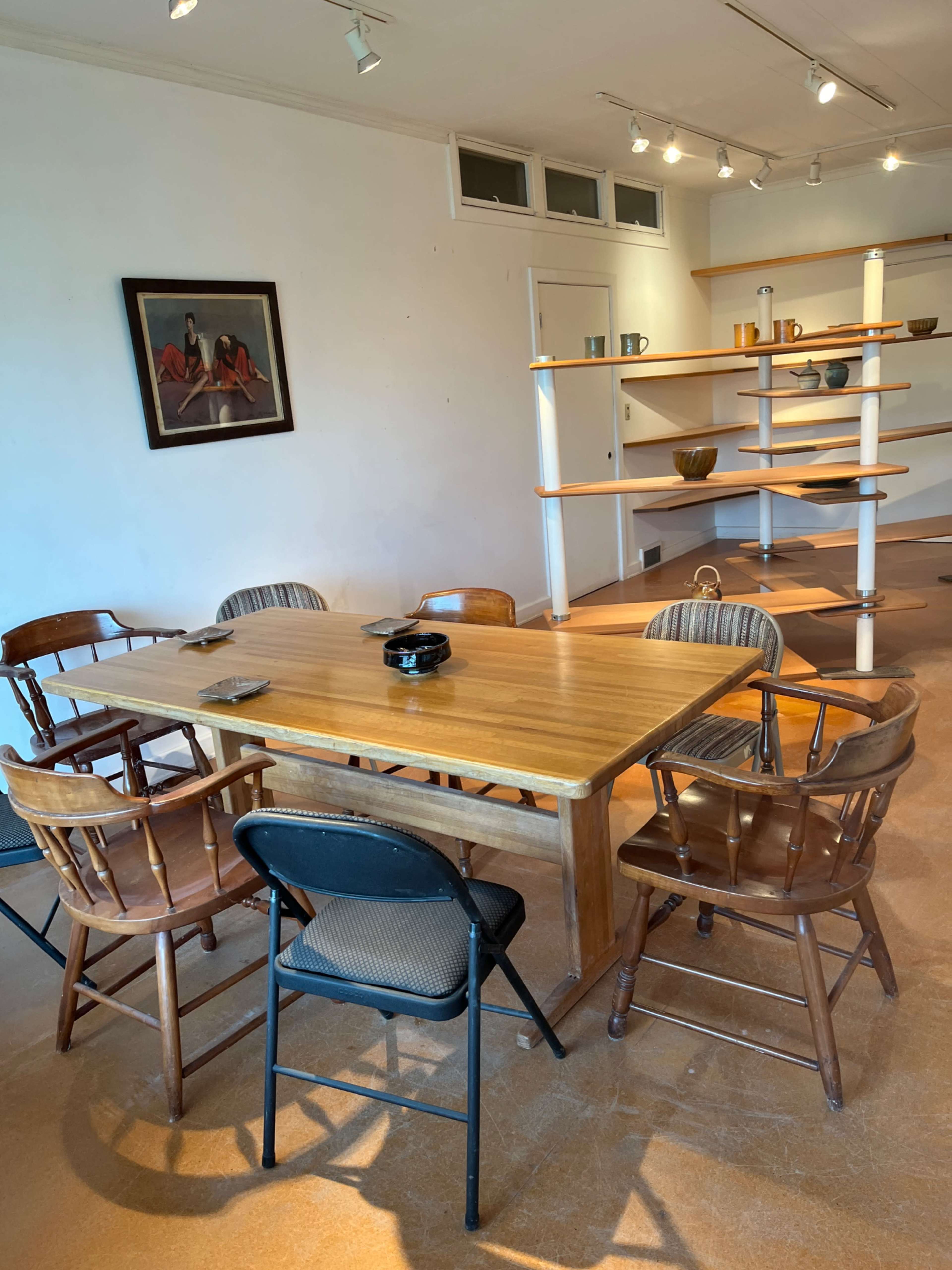 A wooden table surrounded by various chairs is set in a room with a framed painting and a shelf displaying items.