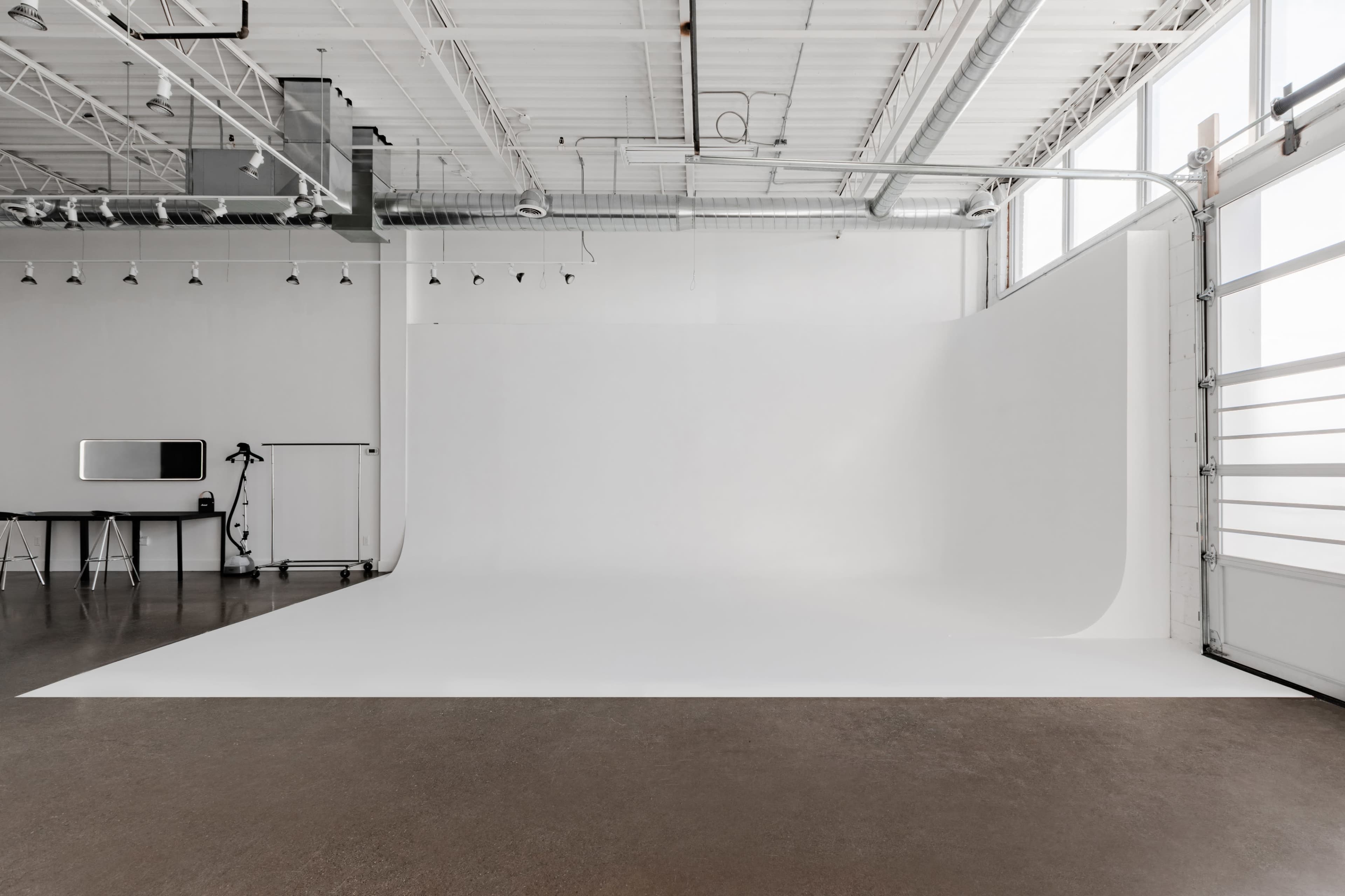 An empty photography studio with a white backdrop, a black table, and a tripod on a polished concrete floor.