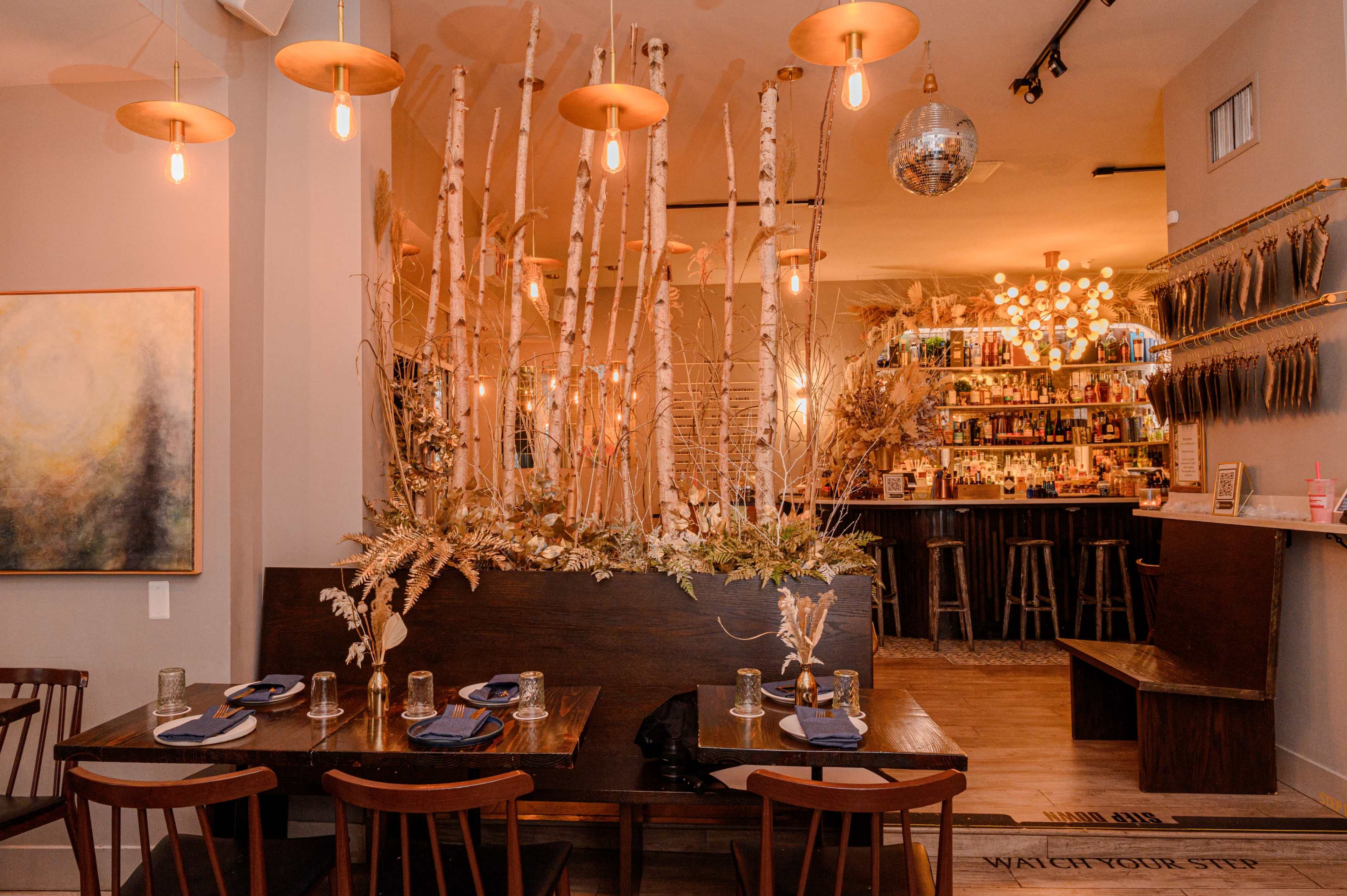 The image shows a stylish restaurant interior featuring wooden tables, hanging lights, and decorative branches, with a bar in the background.
