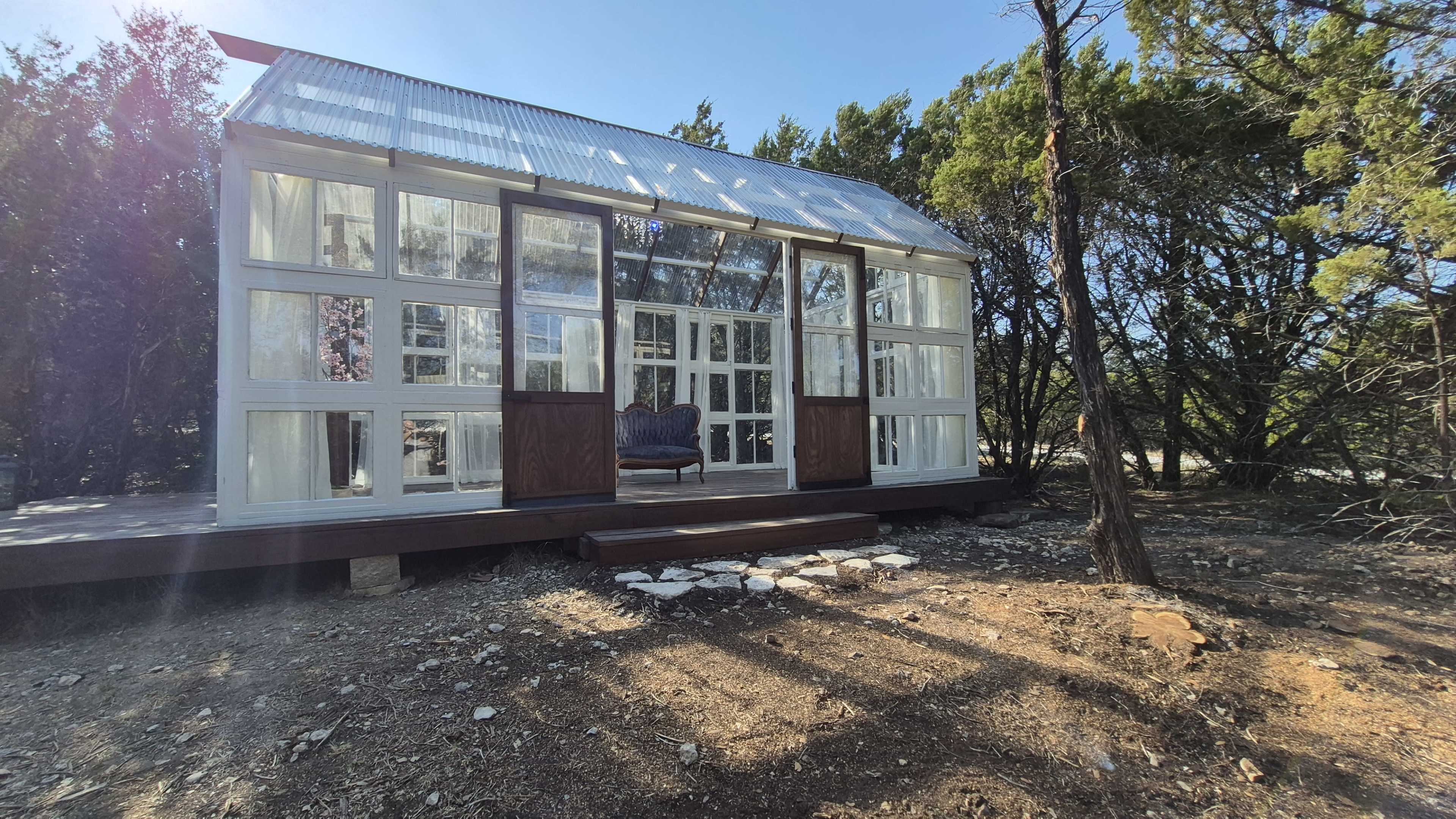 The image shows a modern glass-enclosed house with a wooden deck situated among trees.