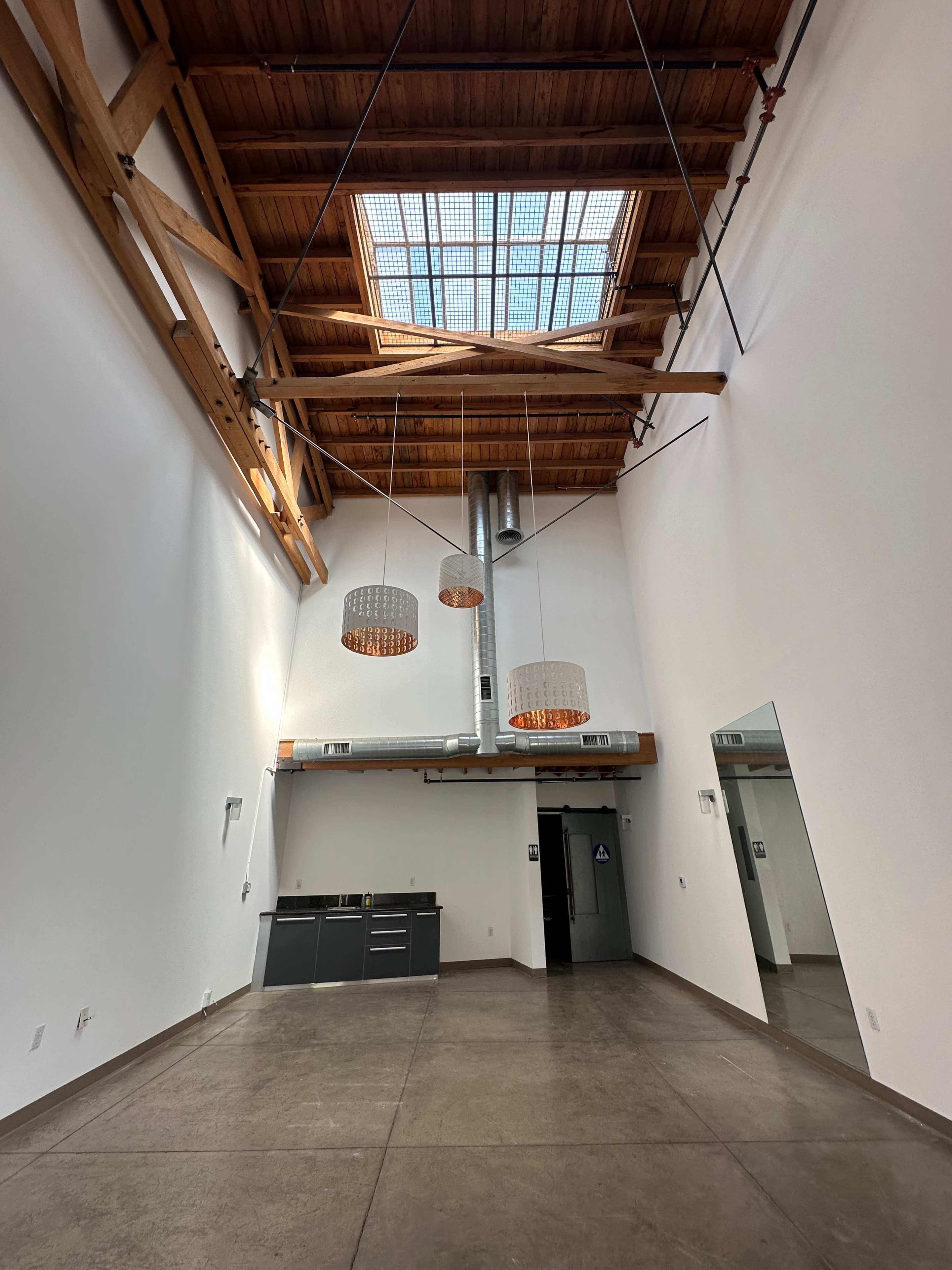The image shows an industrial-style room with high wooden ceilings, three hanging light fixtures, and a kitchen area along one wall.