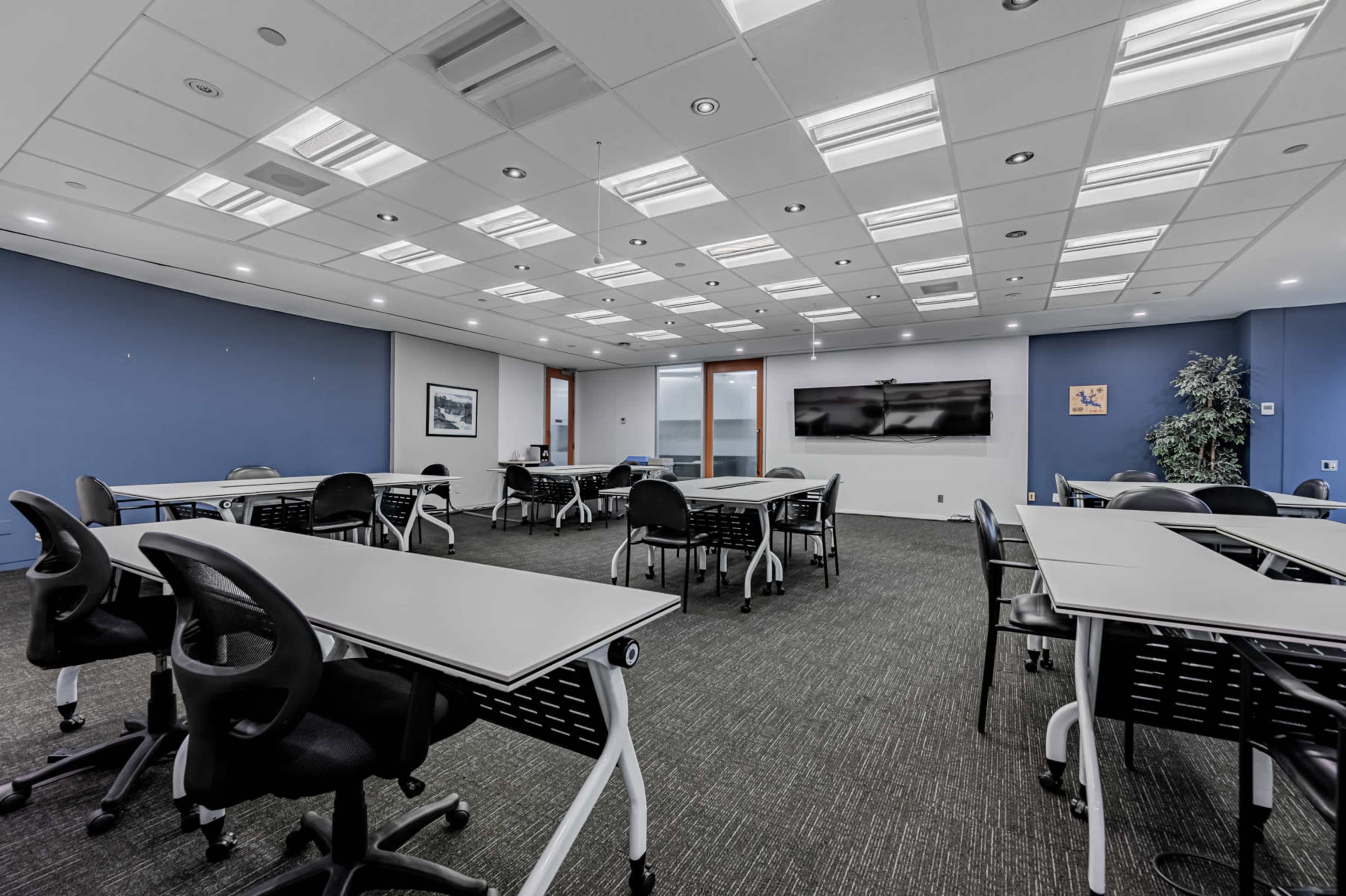 A modern classroom features several empty tables and chairs, with a large screen on one wall and blue accents.