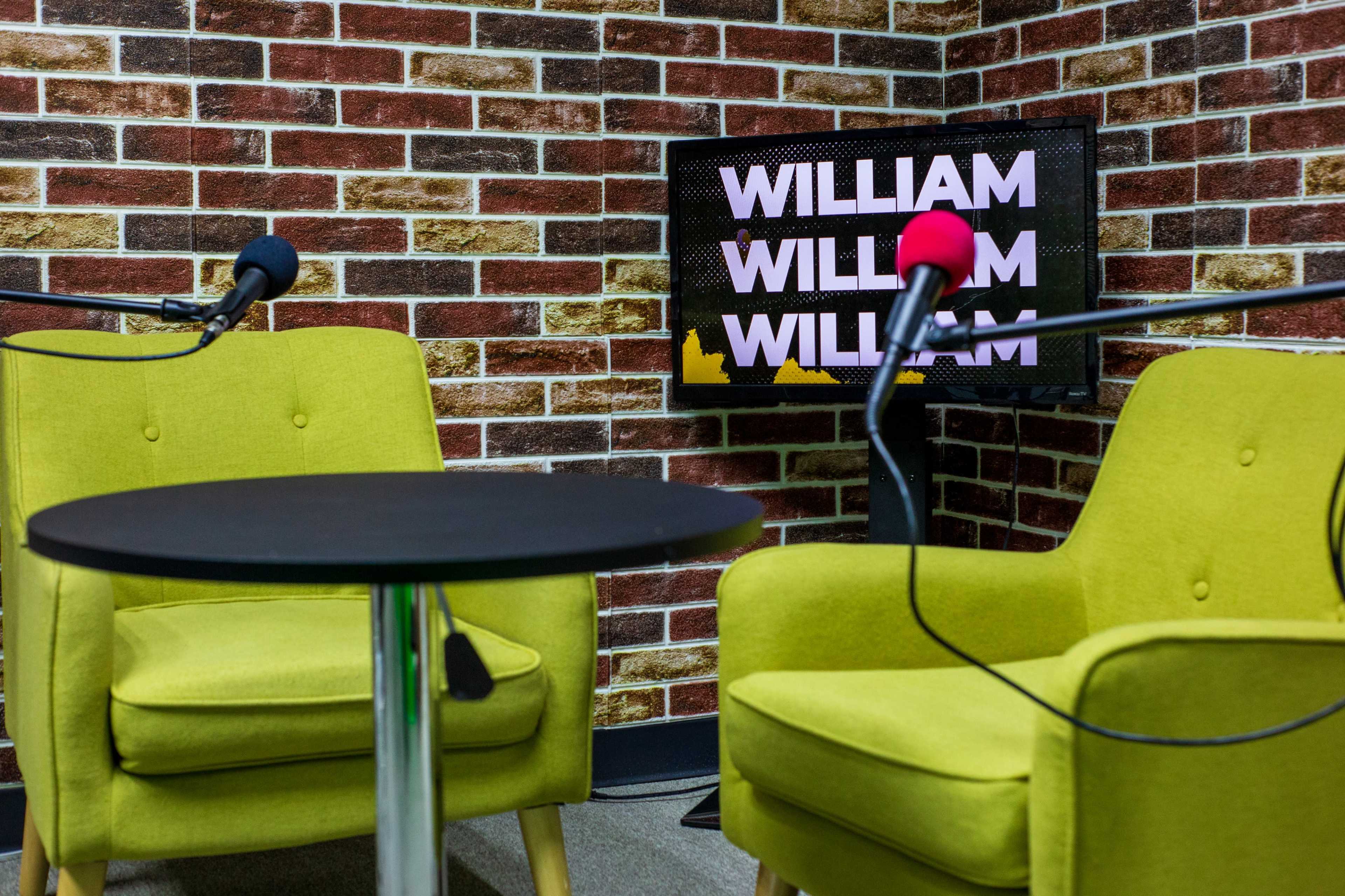 Two green chairs facing a black table with a microphone on each side, set against a brick-patterned wall and a monitor displaying the name "WILLIAM."