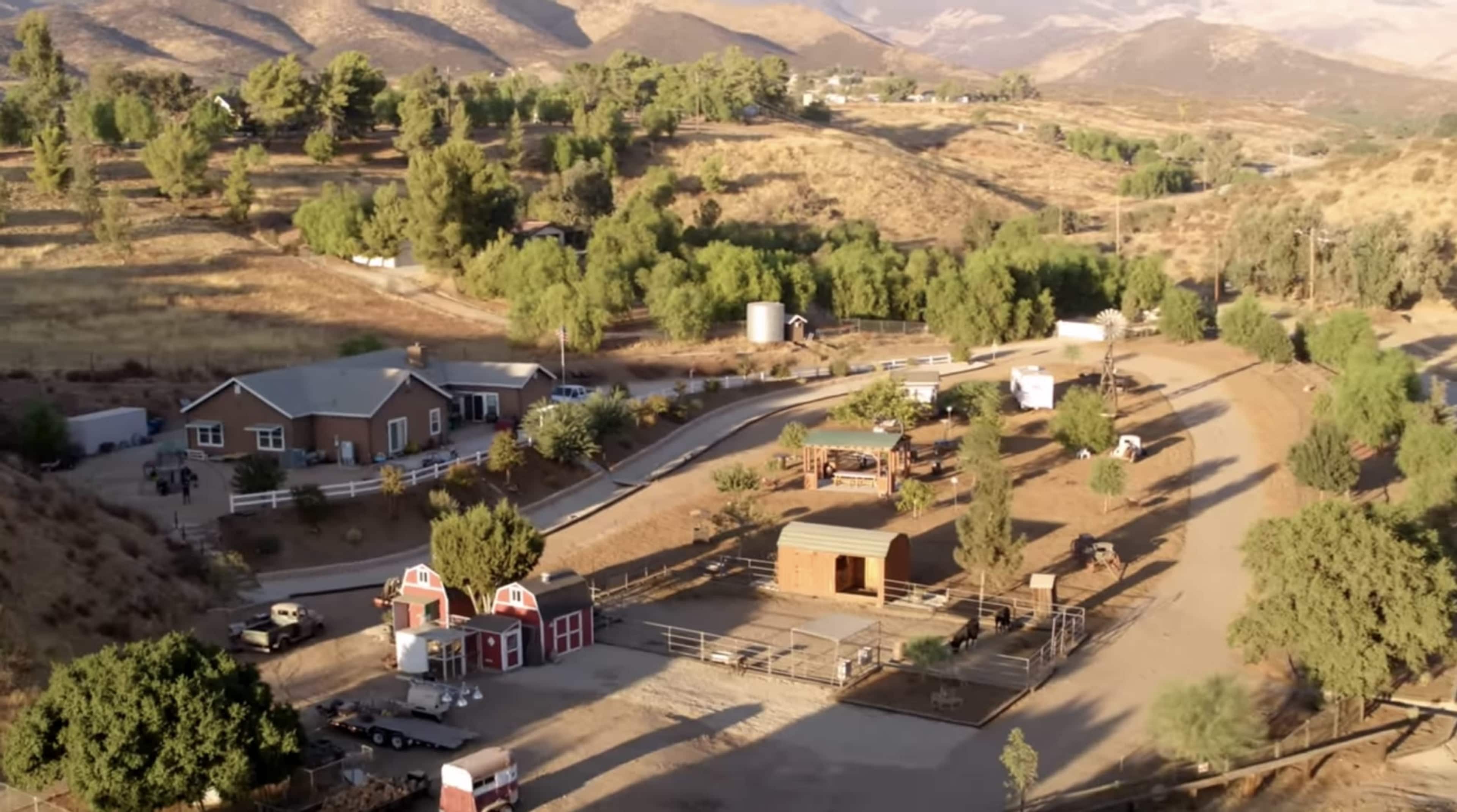 The image shows a rural landscape featuring a cluster of houses, a barn, and several paddocks surrounded by hills and trees.