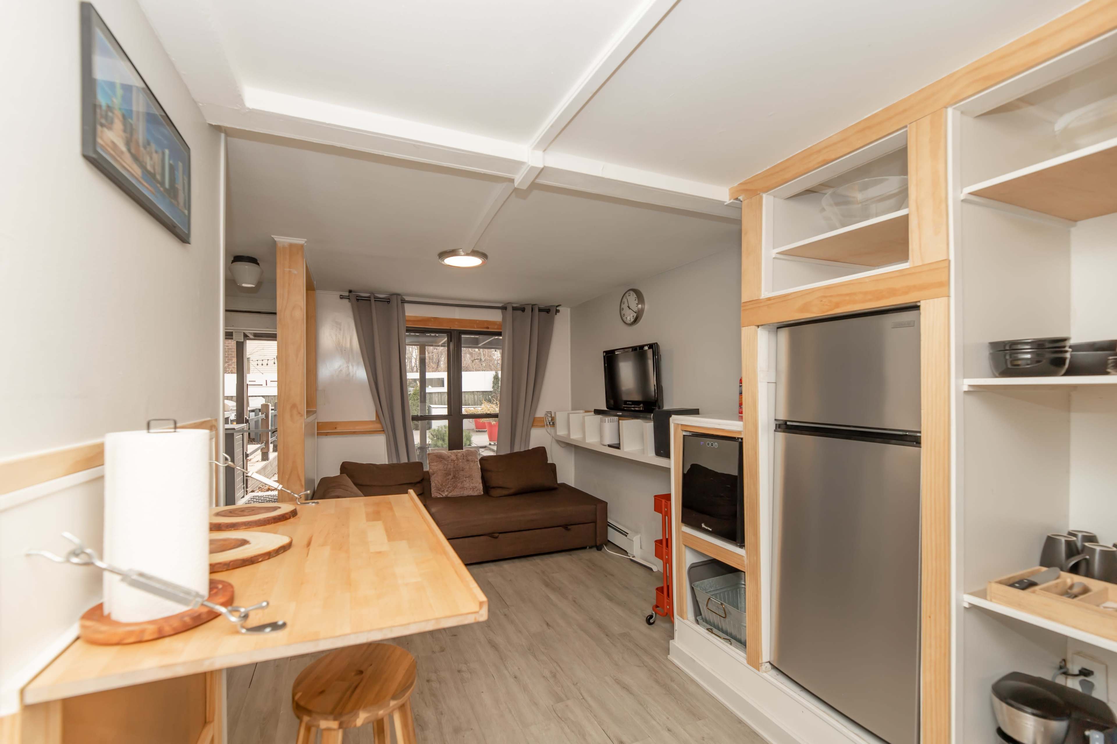 The image shows a compact living space featuring a kitchen area with a wooden table and stools, a couch, shelves, and a refrigerator.