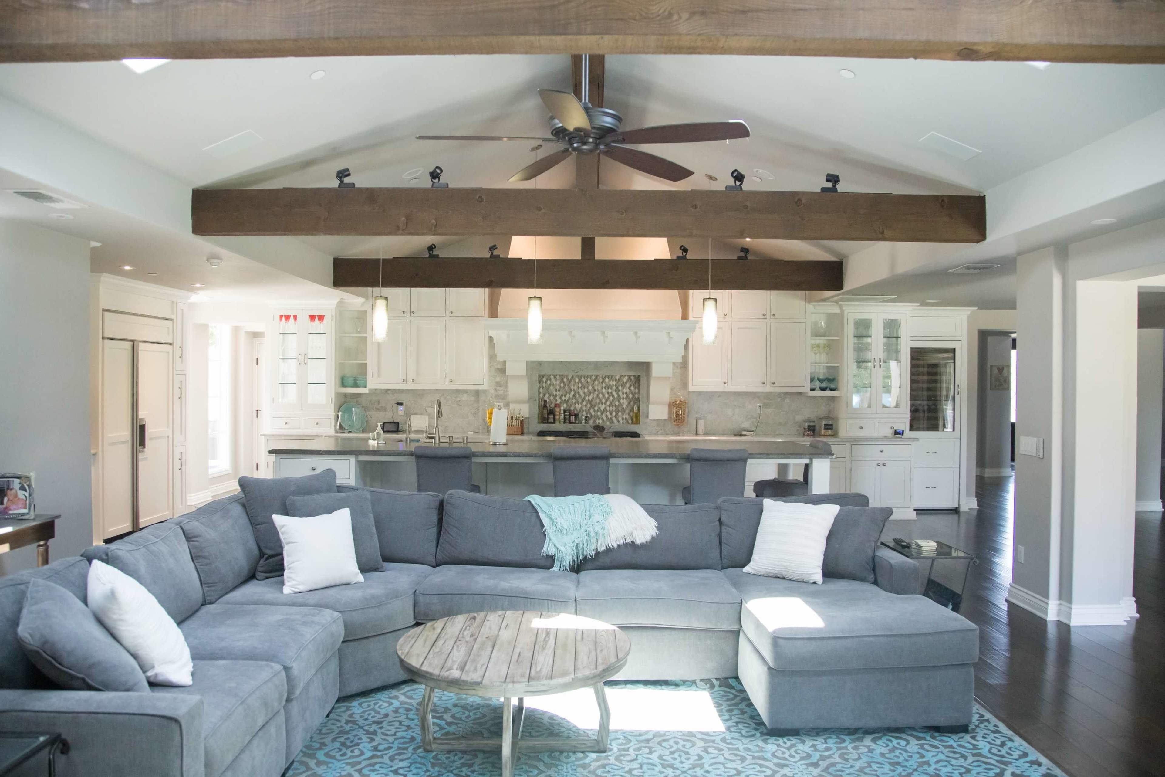 A spacious living room features a large gray sectional sofa, a round wooden coffee table, and exposed wooden beams on the ceiling, with a view of a modern kitchen in the background.