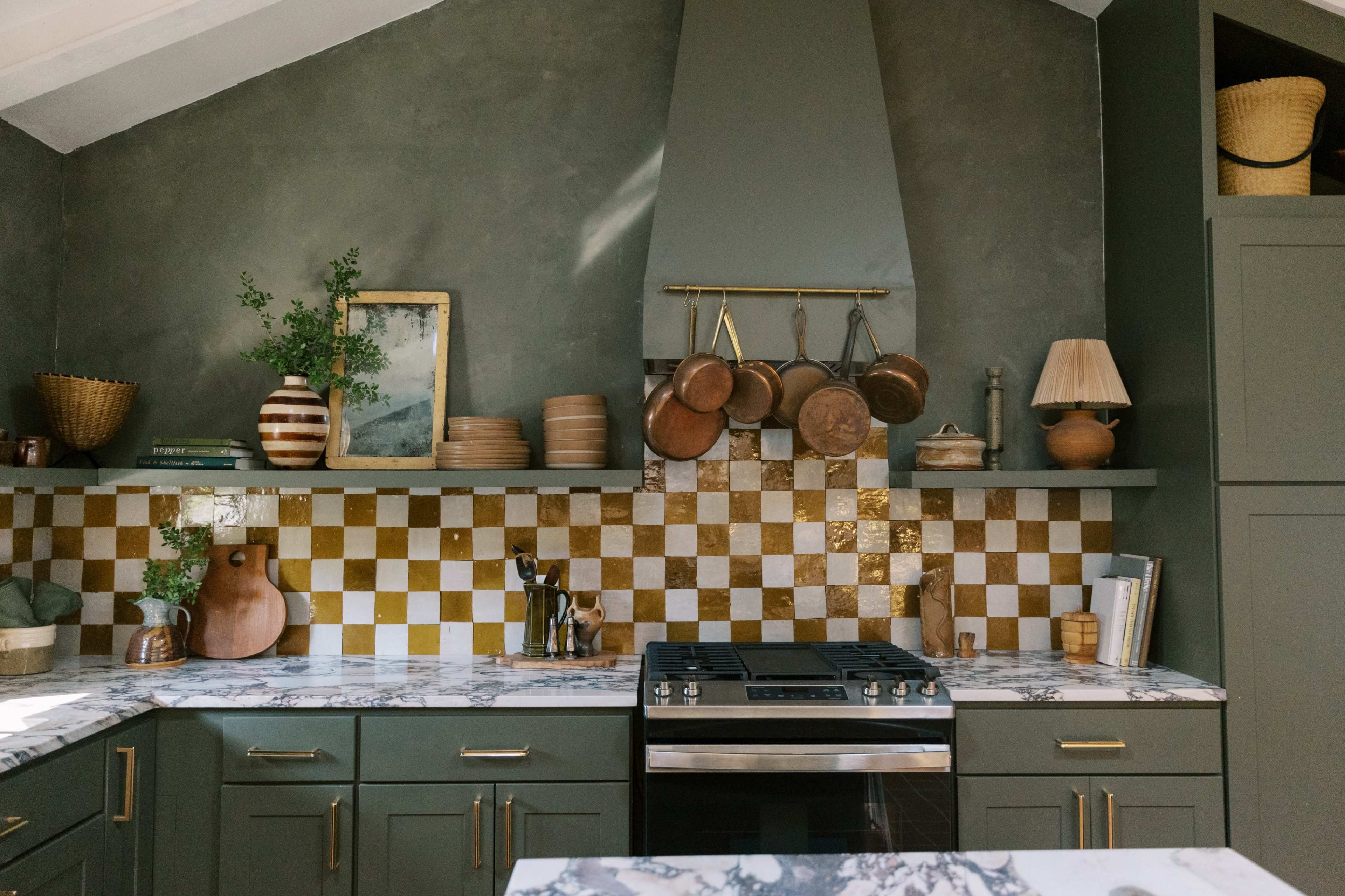 A modern kitchen features green cabinetry, a marble countertop, a checkered gold and white backsplash, and hanging copper pots.