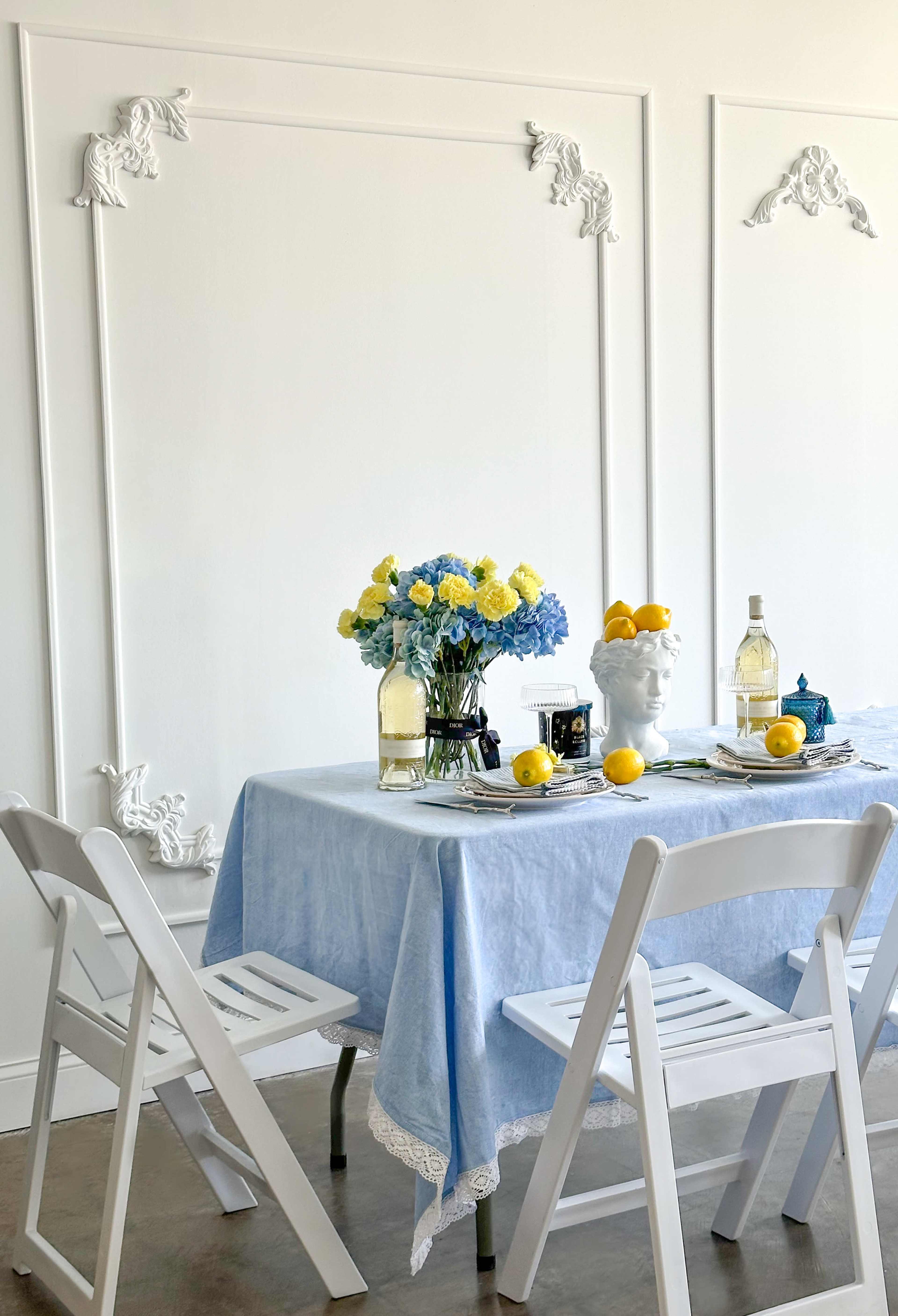 A dining table is set with a light blue tablecloth, featuring a floral arrangement, a bottle of wine, a decorative bowl of lemons, and several empty chairs around it.