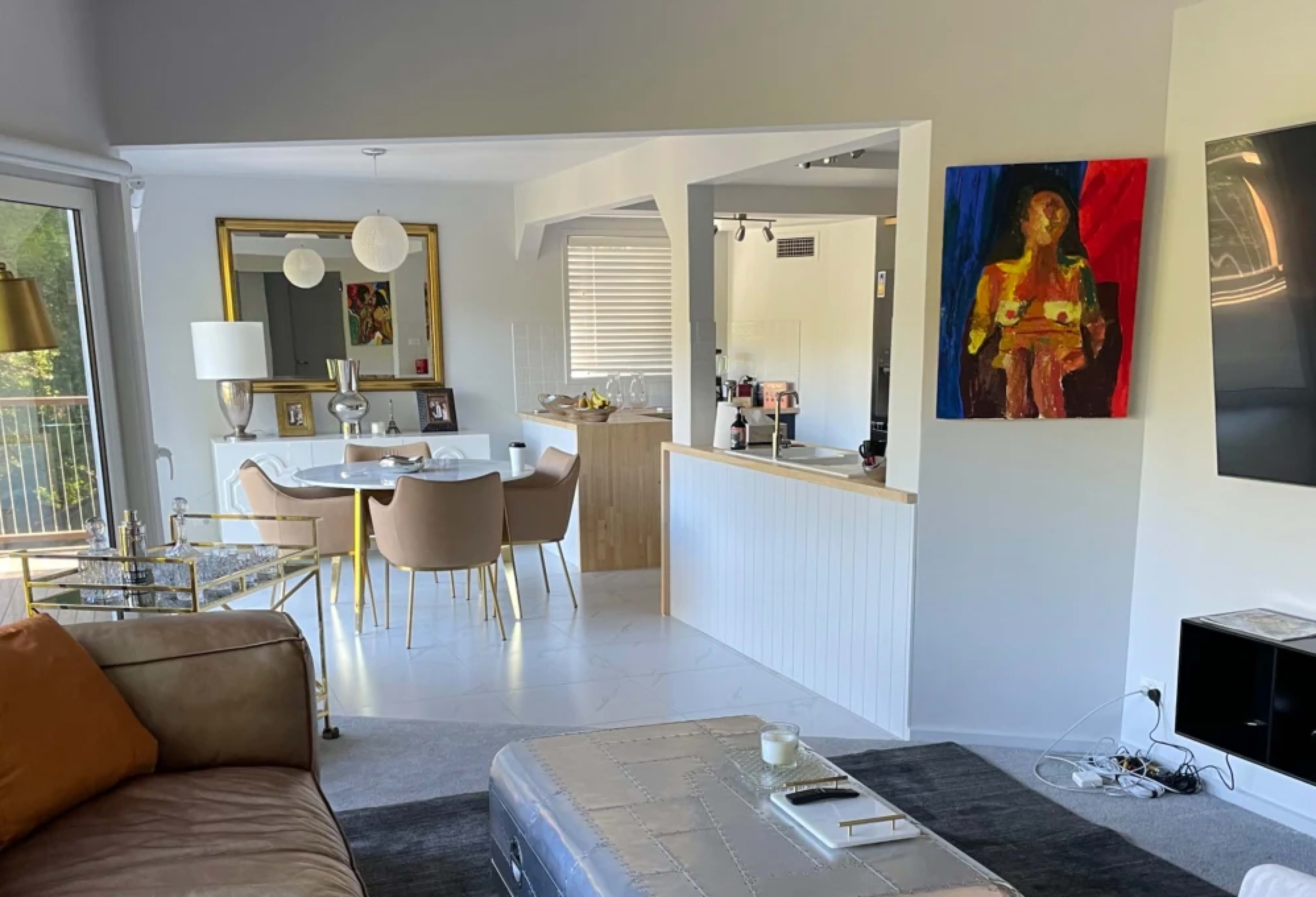 A living room with beige furniture transitions into a dining area featuring a round table and light-colored chairs, adjacent to a kitchen with a large mirror and artwork on the walls.