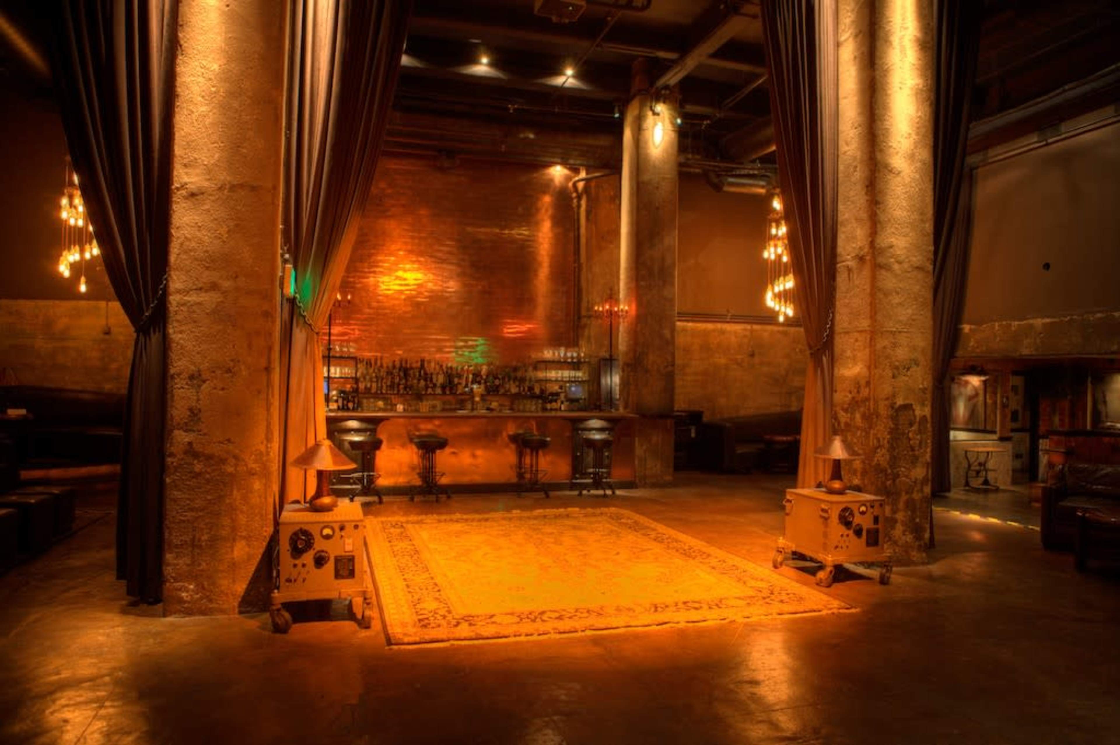 The image depicts a dimly lit bar area with exposed brick walls, wooden flooring, and a prominent illuminated rug in the center, surrounded by bar stools and decorative lighting.