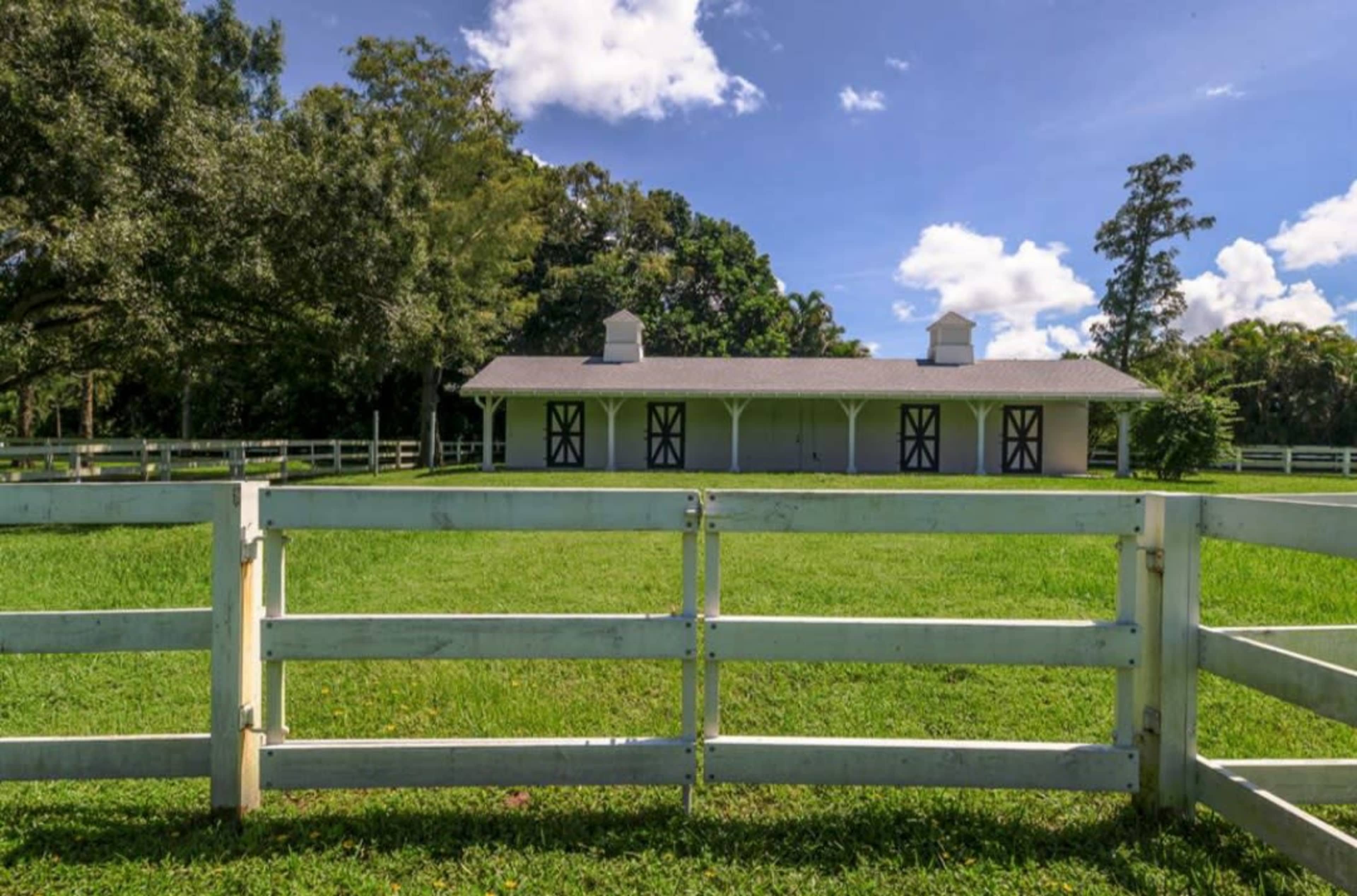 A white fence surrounds a well-maintained green field with a house featuring two cupolas in the background.