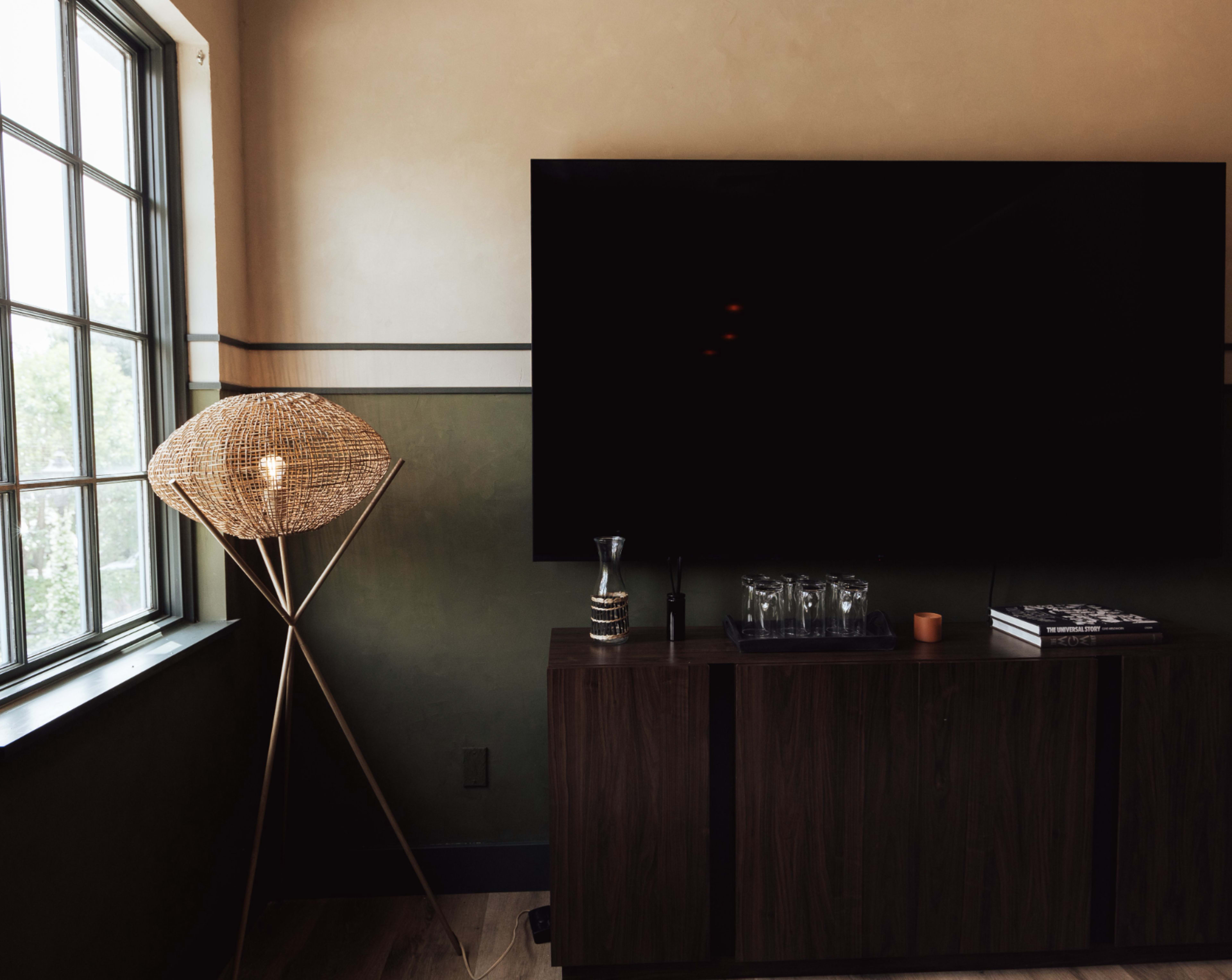 A modern living room features a large black television mounted on the wall next to a wooden cabinet and a floor lamp with a woven shade.