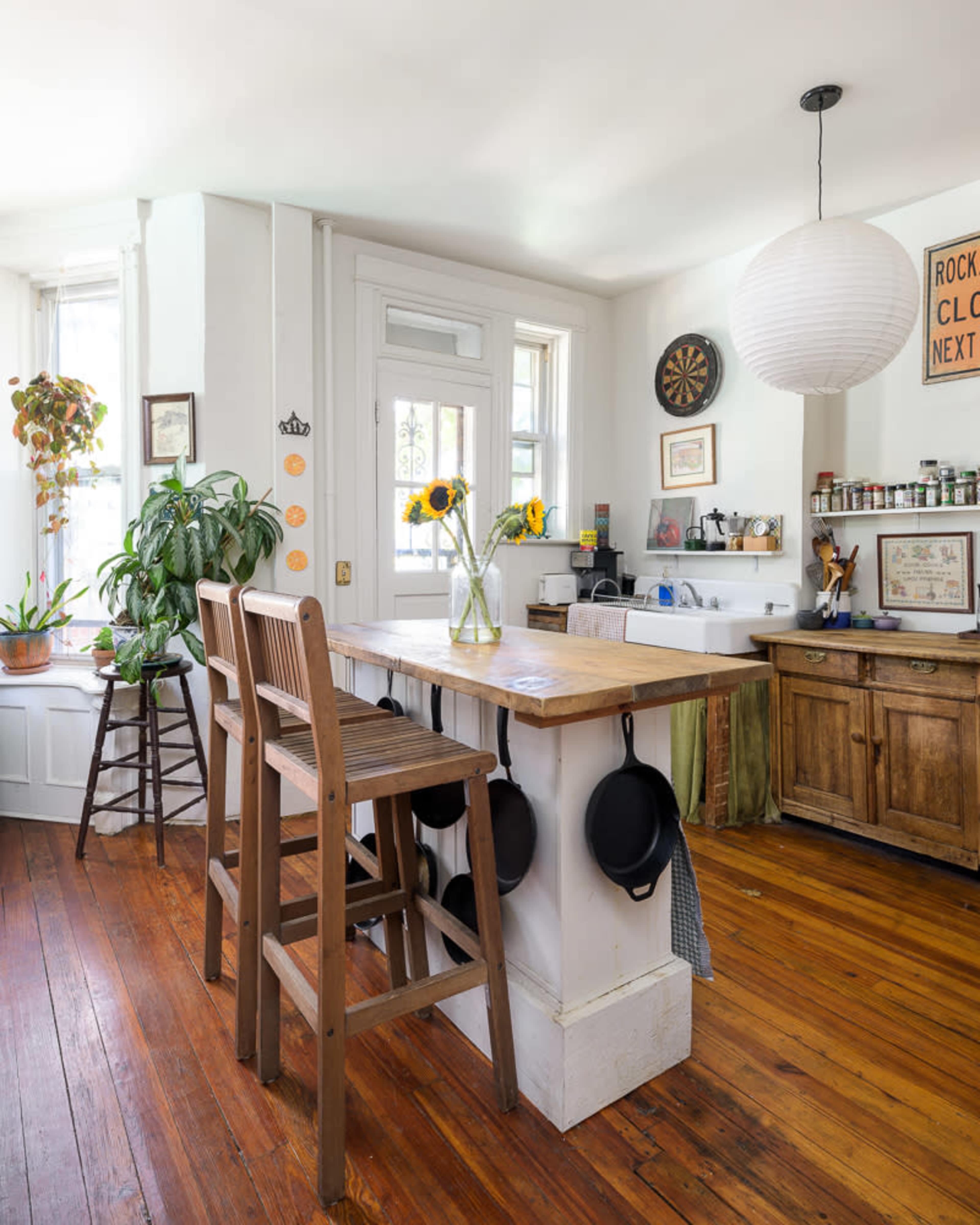 The image shows a cozy kitchen featuring a wooden island with high stools, surrounded by potted plants and natural light streaming through windows.