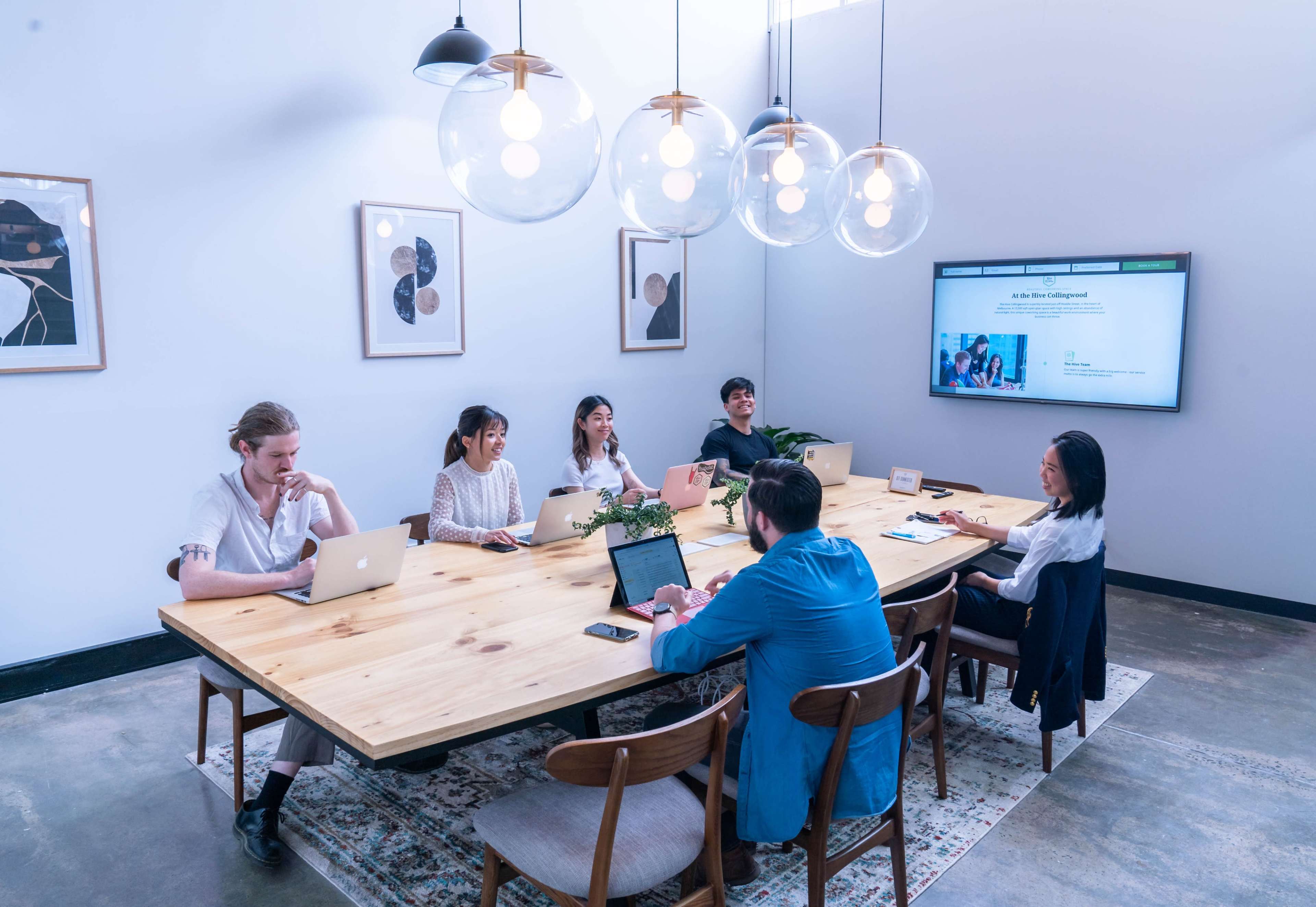 A group of six people sit around a large wooden conference table with laptops, engaged in a meeting in a well-lit room.