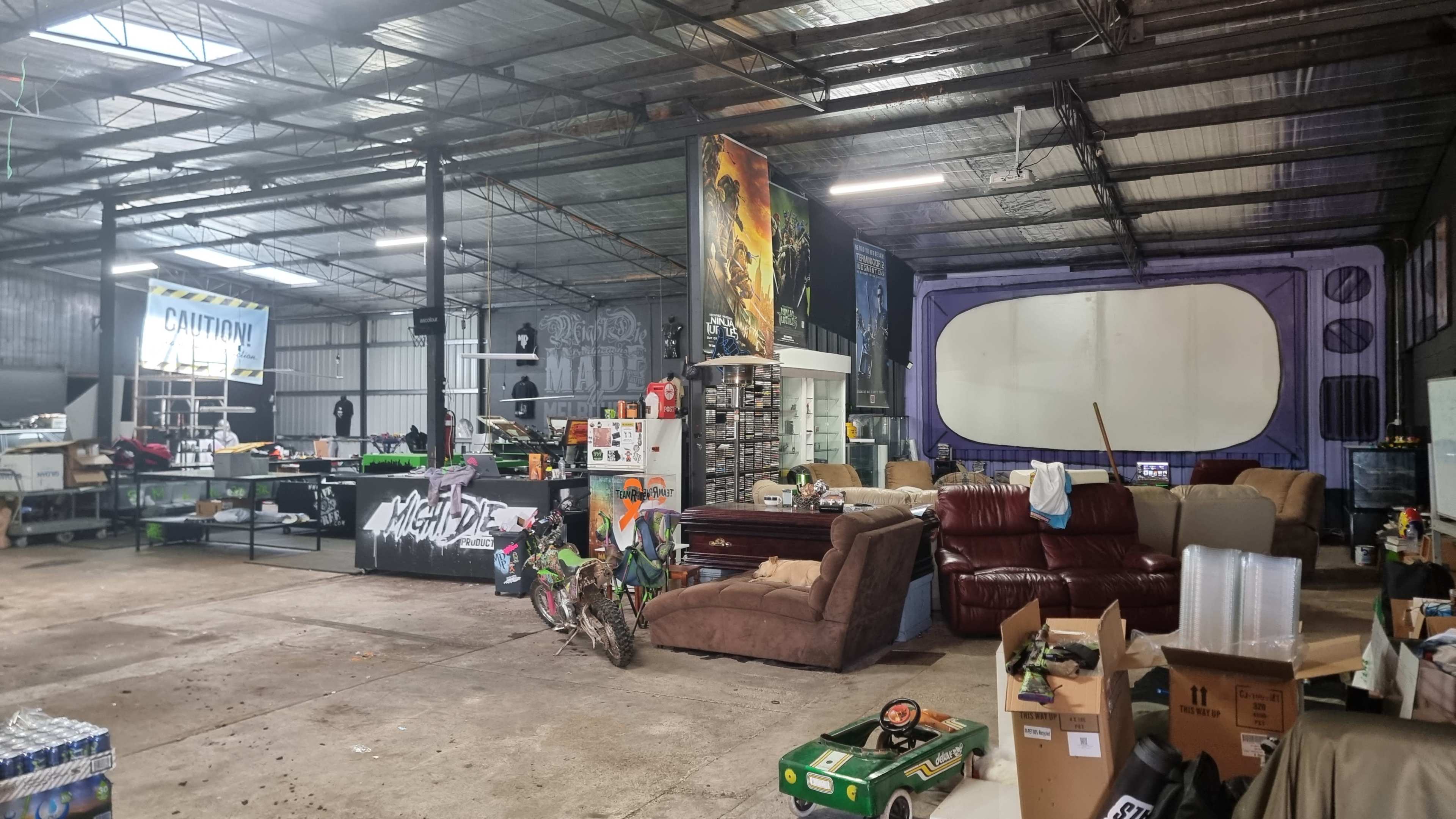 An industrial warehouse space filled with various furniture, equipment, and boxes, with a dirt floor and exposed metal beams.