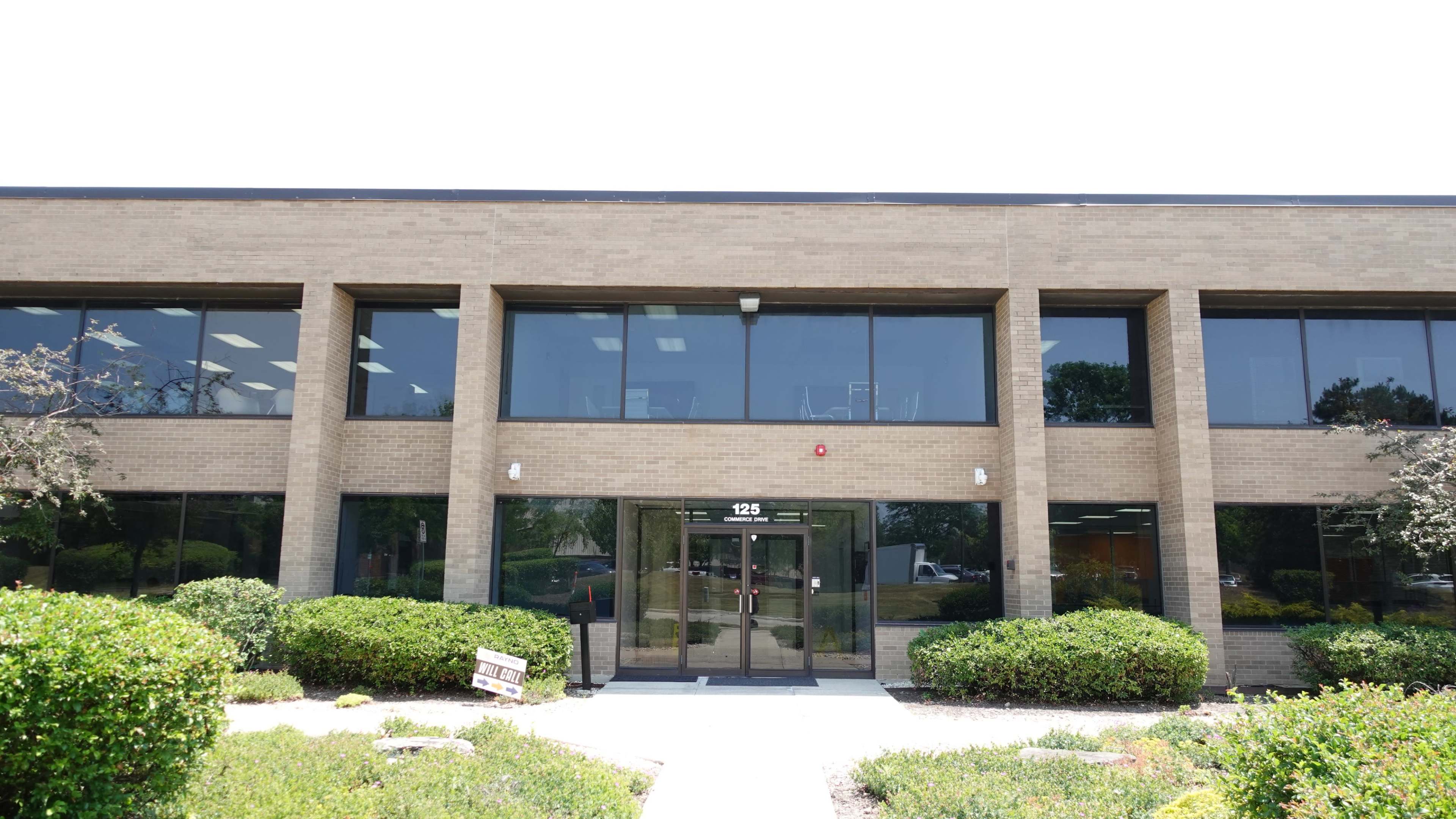 The image shows a modern two-story office building with large windows and a glass entrance, surrounded by neatly trimmed shrubs and landscaping.