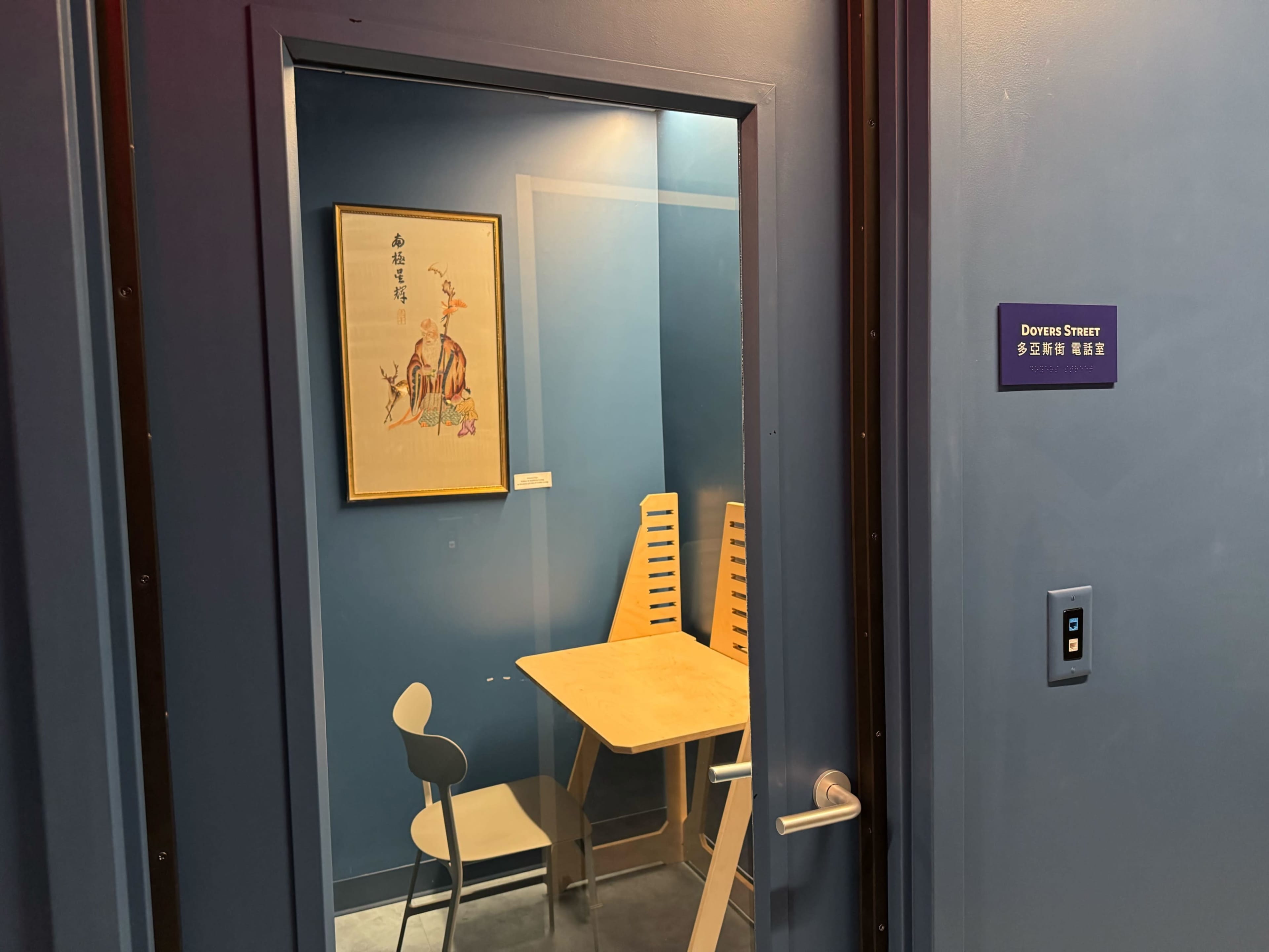 The image shows a small room with a blue wall, a wooden table and chairs, and a framed artwork hanging on the wall.