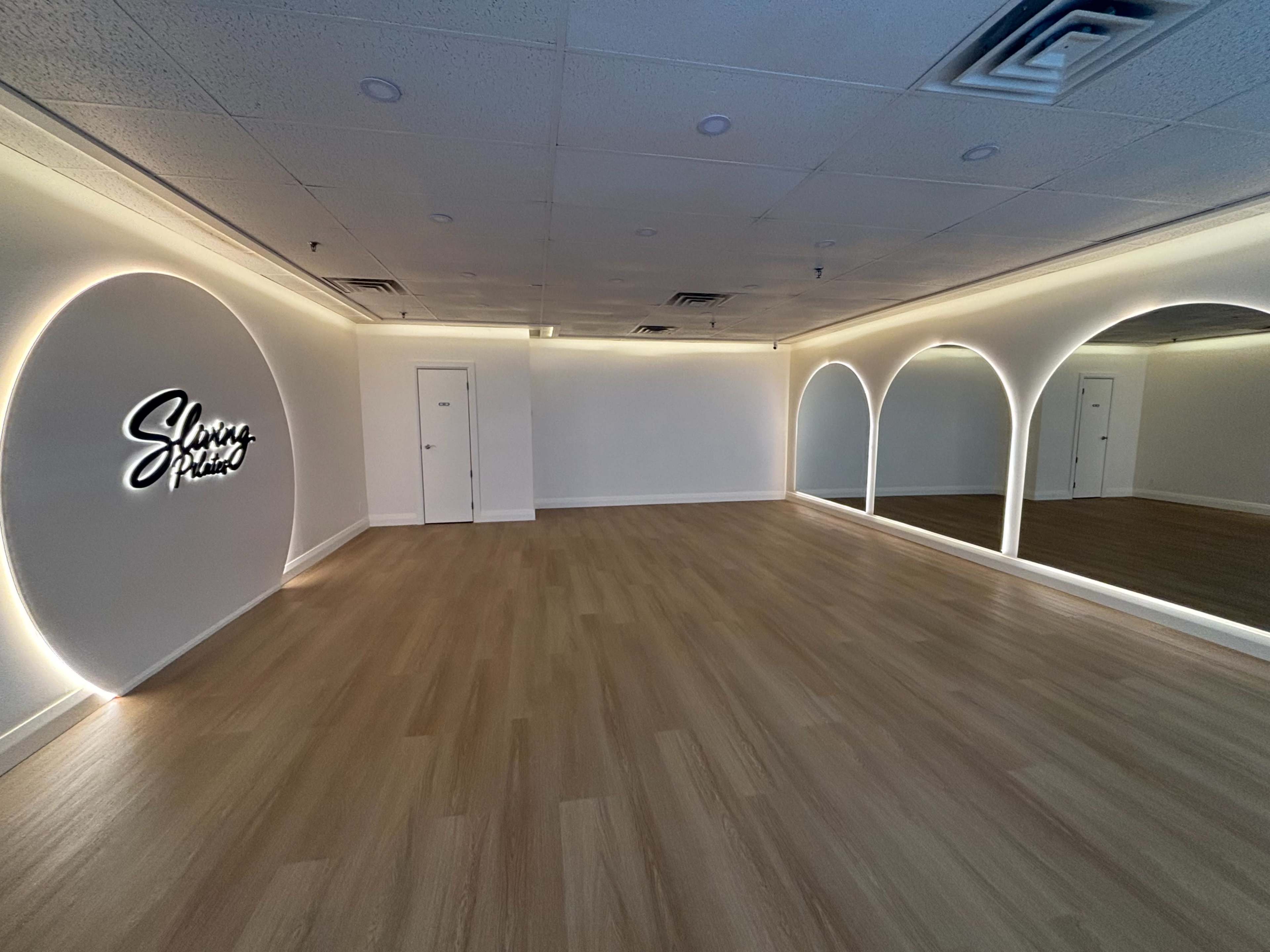 The image shows an empty, well-lit studio space with wooden flooring, softly illuminated archways, and a wall featuring the name "Shiny Prism" in stylized lettering.