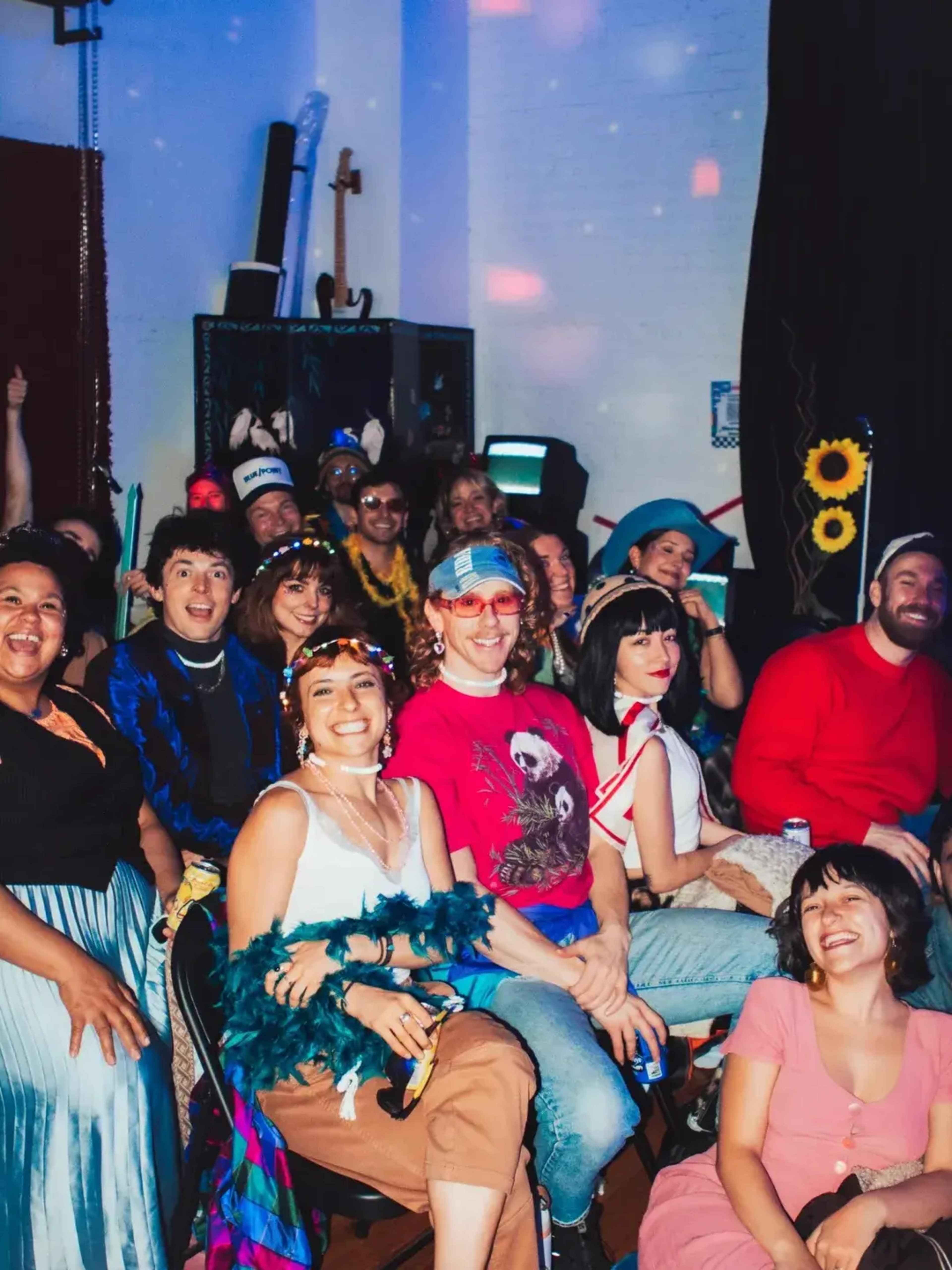 A diverse group of people, dressed in colorful outfits, smiles and poses for the camera in a lively indoor setting.