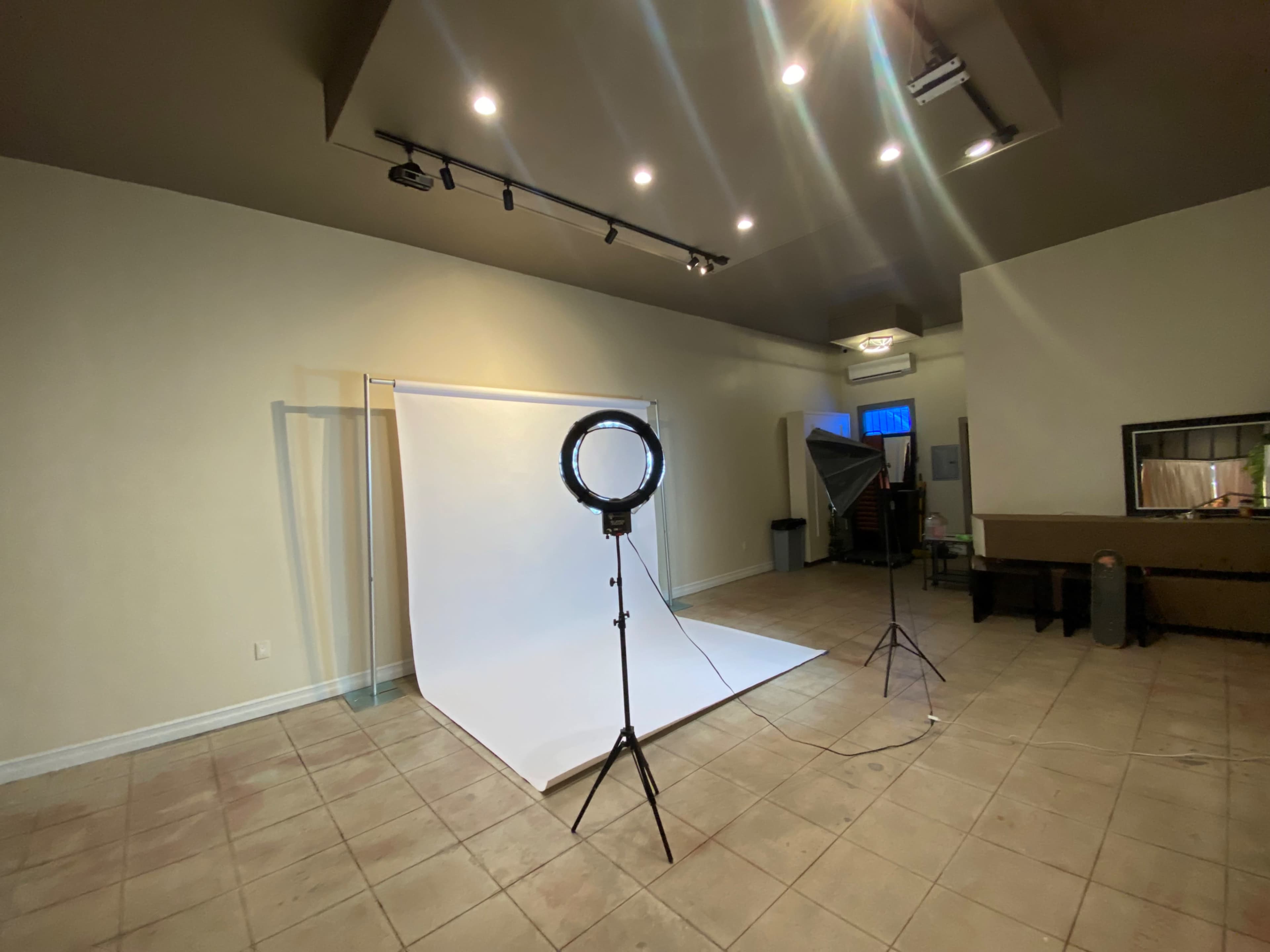 A photography studio is set up with a white backdrop, a ring light on a stand, and several overhead lights.