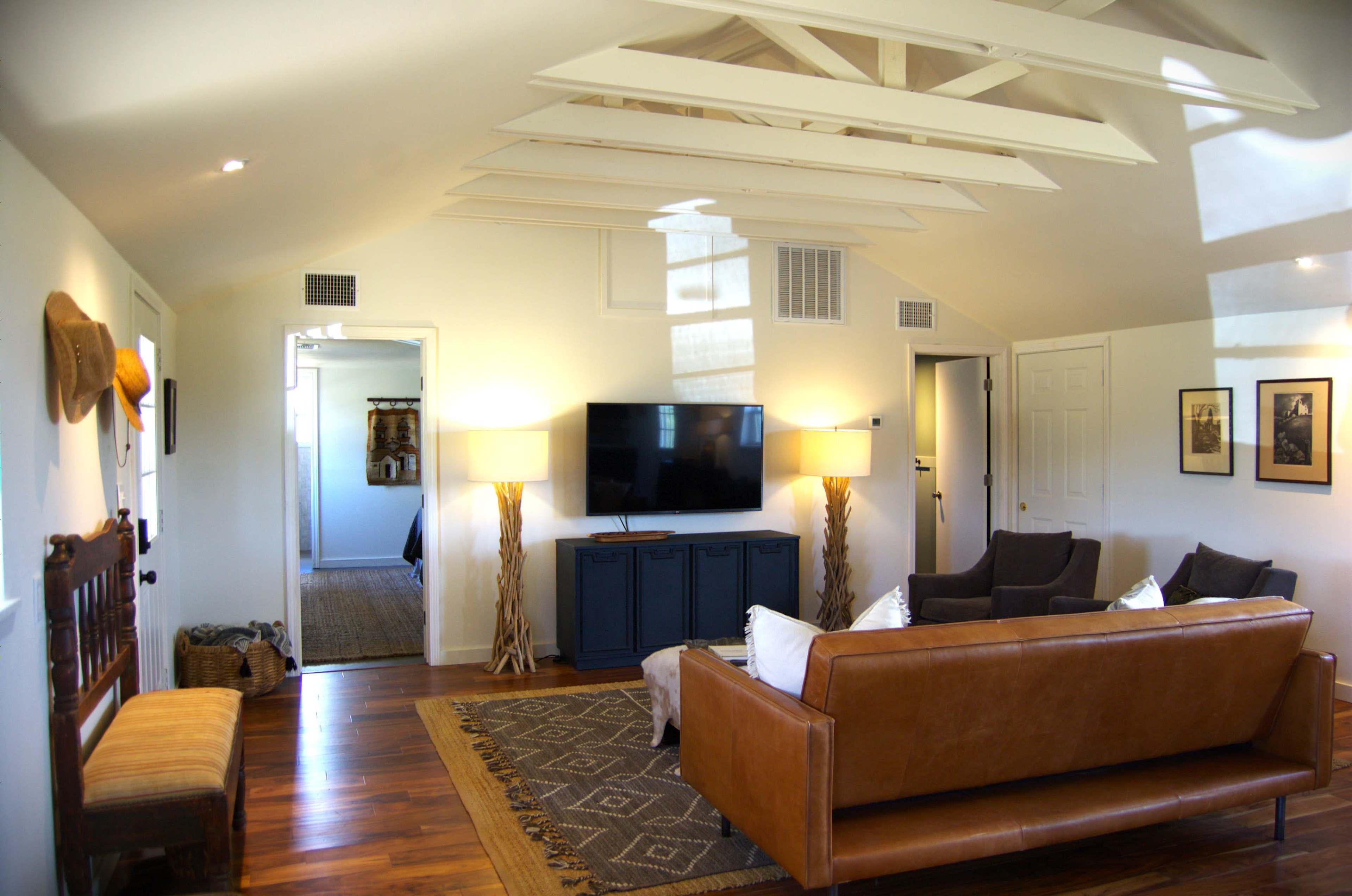 The image shows a spacious living room with a brown leather sofa, two dark chairs, a blue television cabinet, and wooden beams on the ceiling, illuminated by natural light.